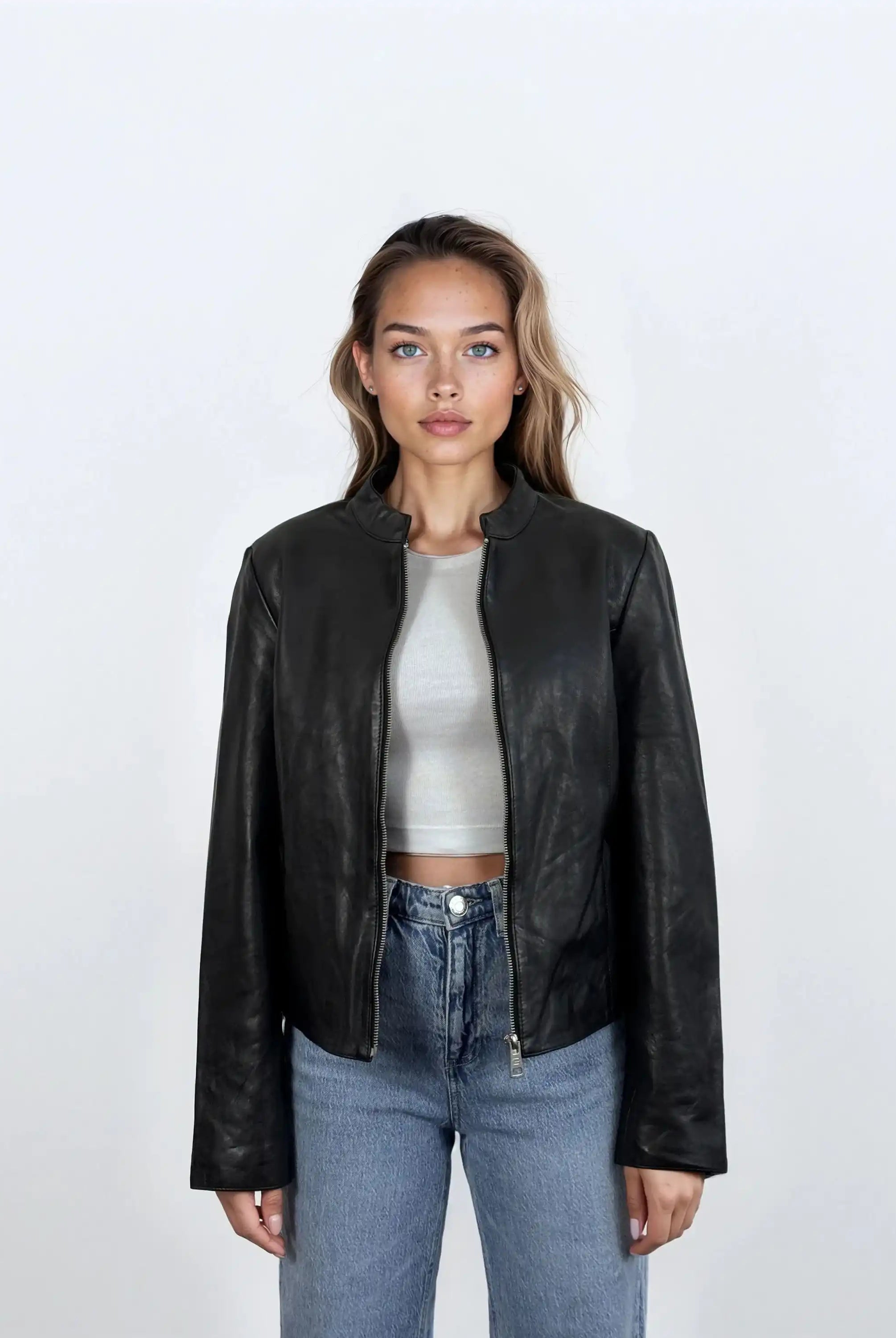 Eine Frau mit langem, gewelltem Haar und neutralem Gesichtsausdruck trägt die trueprodigy Damen Lederjacke aus Schafsleder in Schwarz mit minimalistischem Design über einem hellgrauen Crop-Top und blauen Jeans vor einem schlichten weißen Hintergrund.