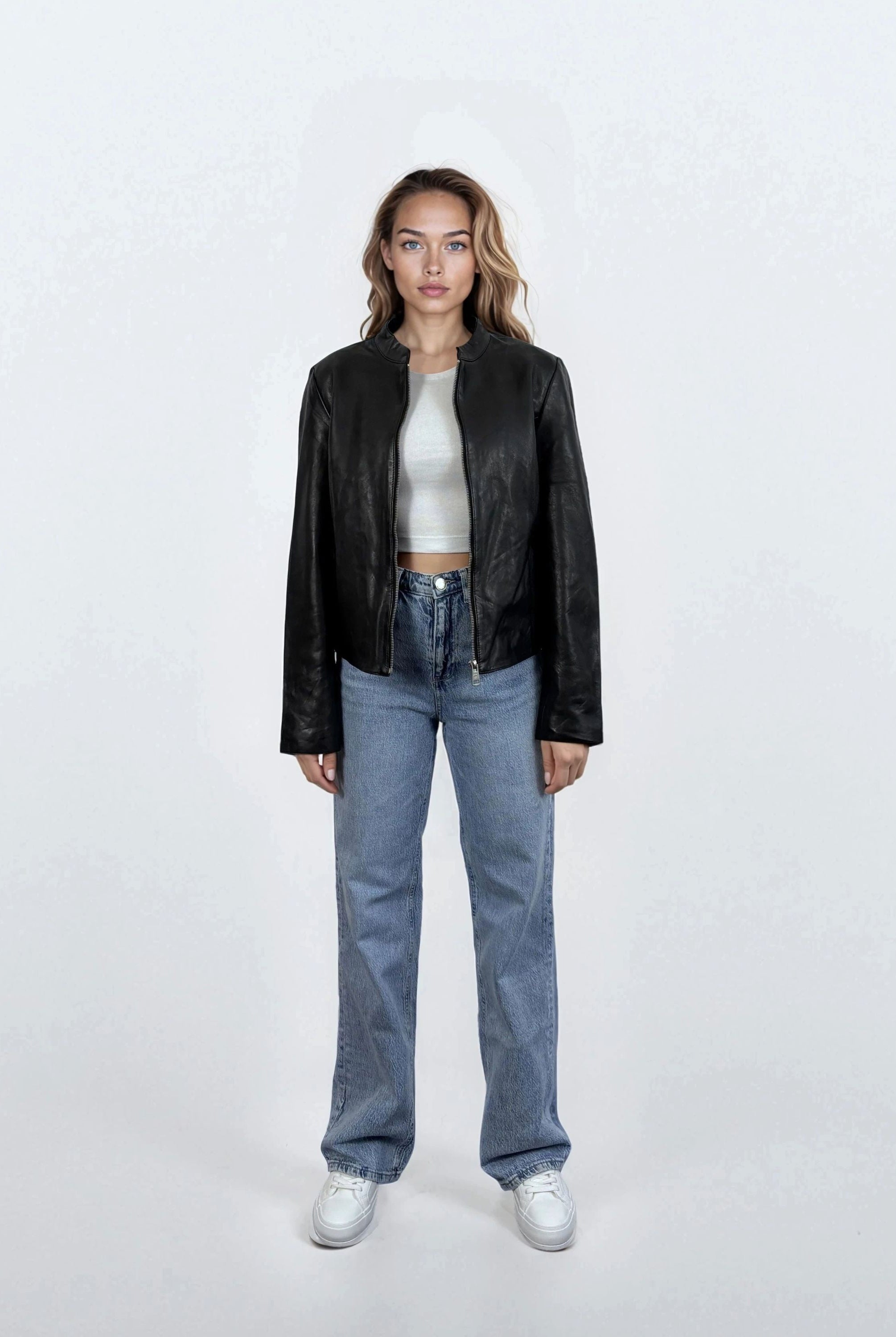 Eine Frau steht mit dem Gesicht nach vorne vor einem weißen Hintergrund und trägt eine trueprodigy Damen Lederjacke aus Schafsleder in Schwarz mit minimalistischem Design, gepaart mit einem weißen Crop-Top, blauen Jeans und weißen Sneakers. Ihre Arme sind an den Seiten verschränkt.