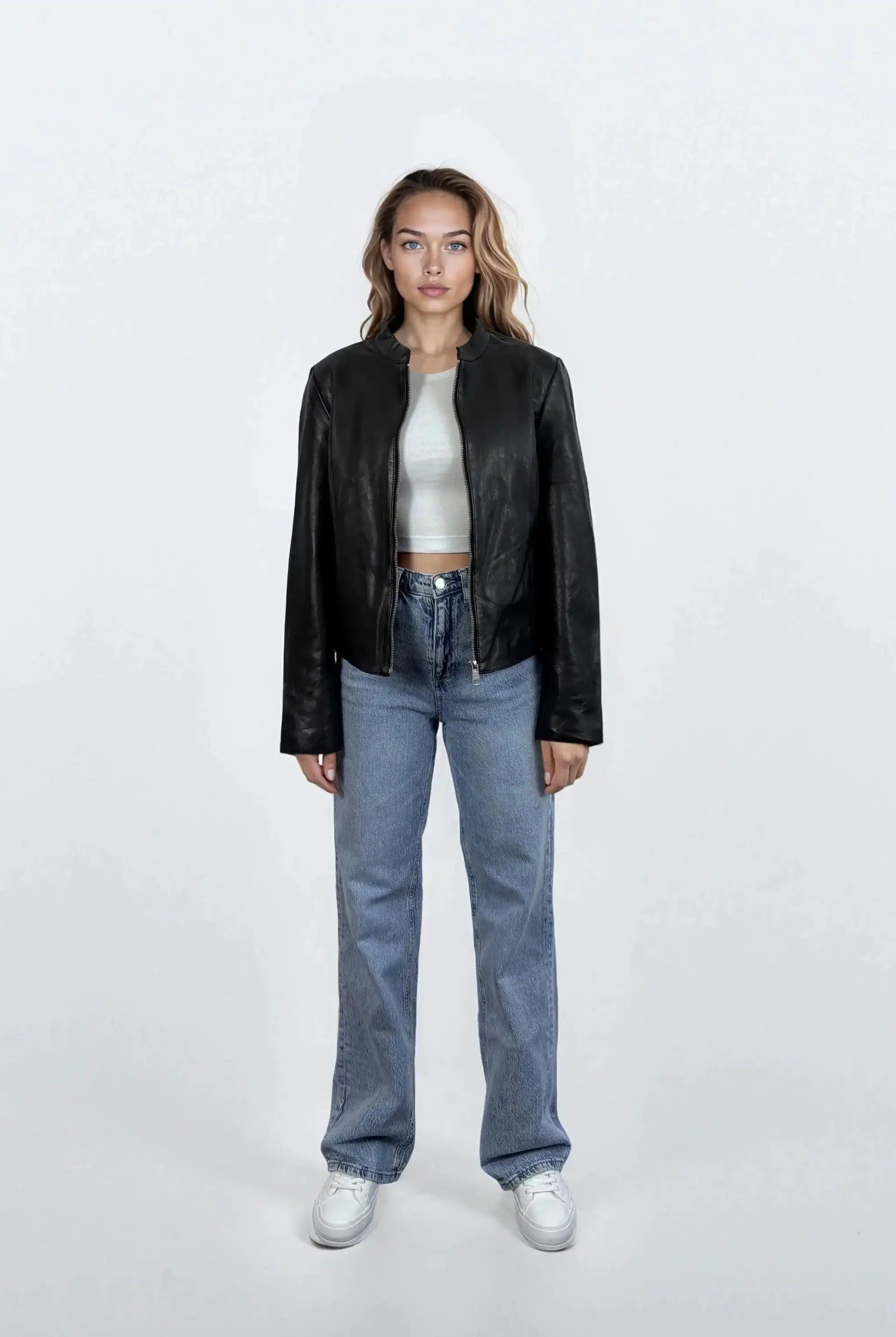 Eine Frau steht mit dem Gesicht nach vorne vor einem weißen Hintergrund und trägt eine trueprodigy Damen Lederjacke aus Schafsleder in Schwarz mit minimalistischem Design, gepaart mit einem weißen Crop-Top, blauen Jeans und weißen Sneakers. Ihre Arme sind an den Seiten verschränkt.