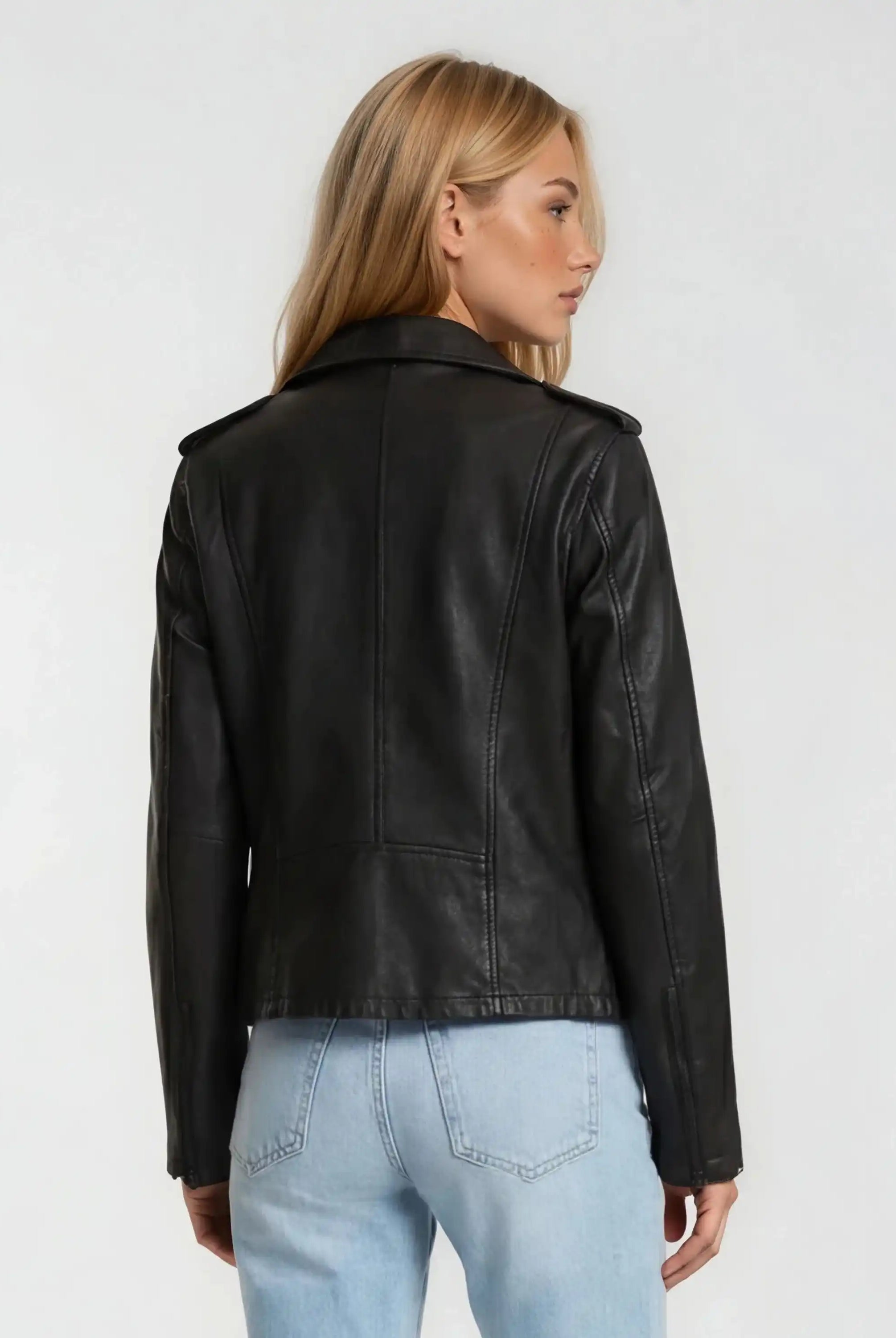 Man sieht eine Frau mit langen blonden Haaren von hinten, die die trueprodigy Damen Lederjacke aus Schafsleder in Schwarz im klassischen Biker-Stil und hellblaue Jeans vor einem schlichten hellen Hintergrund trägt.