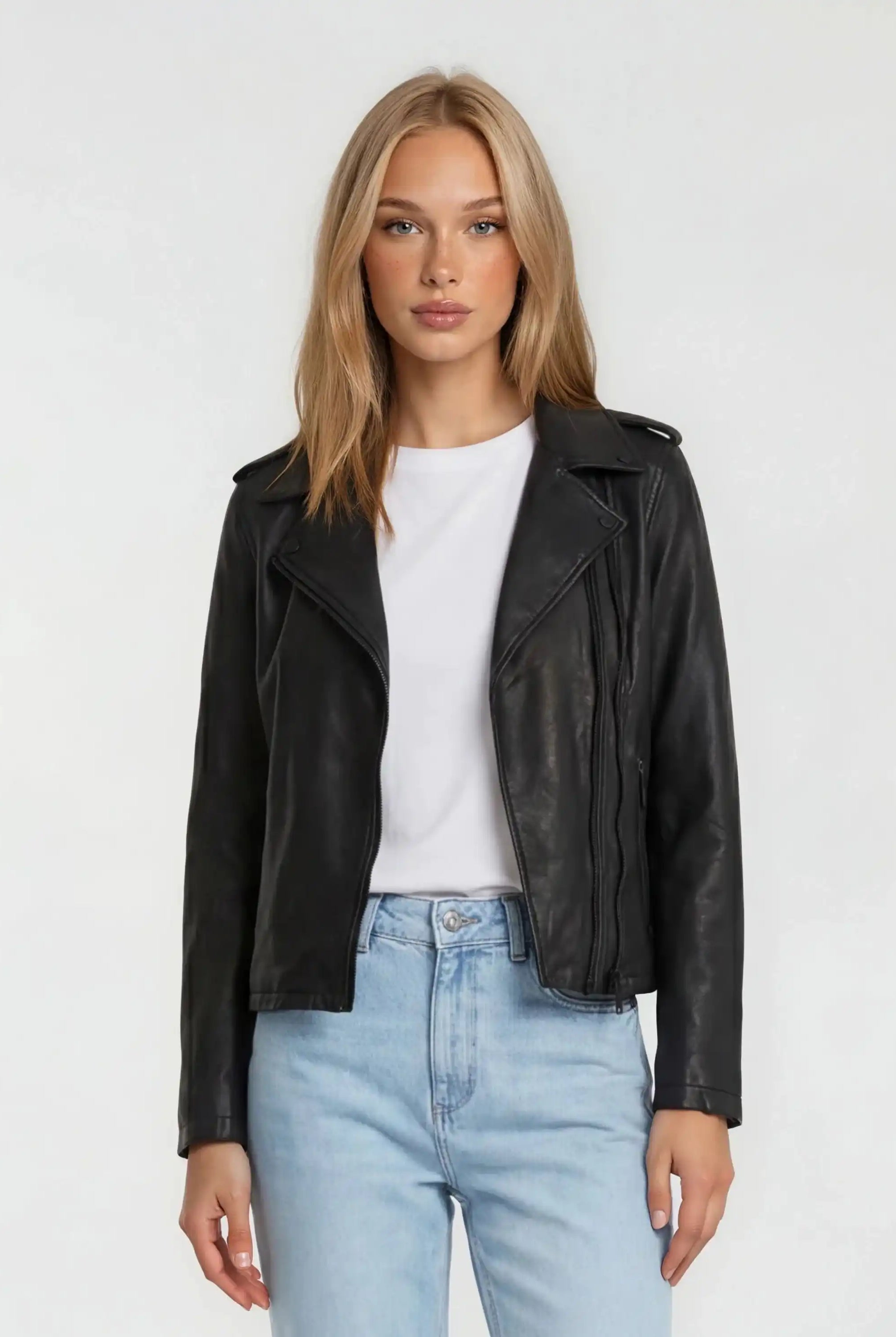 Eine Person mit glattem blondem Haar trägt eine trueprodigy Damen Lederjacke aus Schafsleder in Schwarz im klassischen Biker-Stil über einem weißen T-Shirt und hellblauen Jeans, die vor einem schlichten hellen Hintergrund stehen.