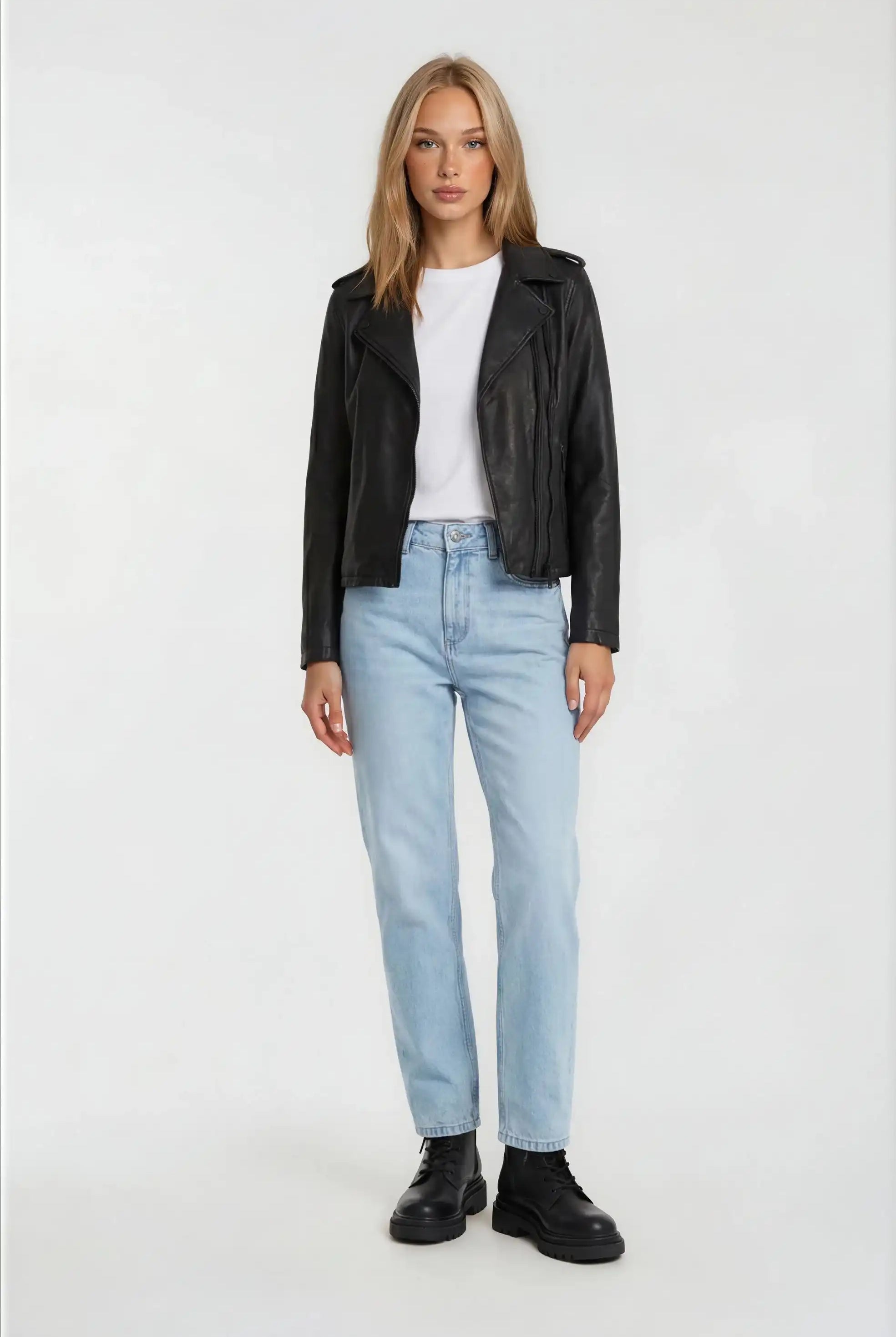 Eine Person steht vor einem weißen Hintergrund und trägt die trueprodigy Damen Lederjacke aus Schafsleder in Schwarz im klassischen Biker-Stil, ein weißes T-Shirt, hellblaue Jeans und schwarze Stiefel.