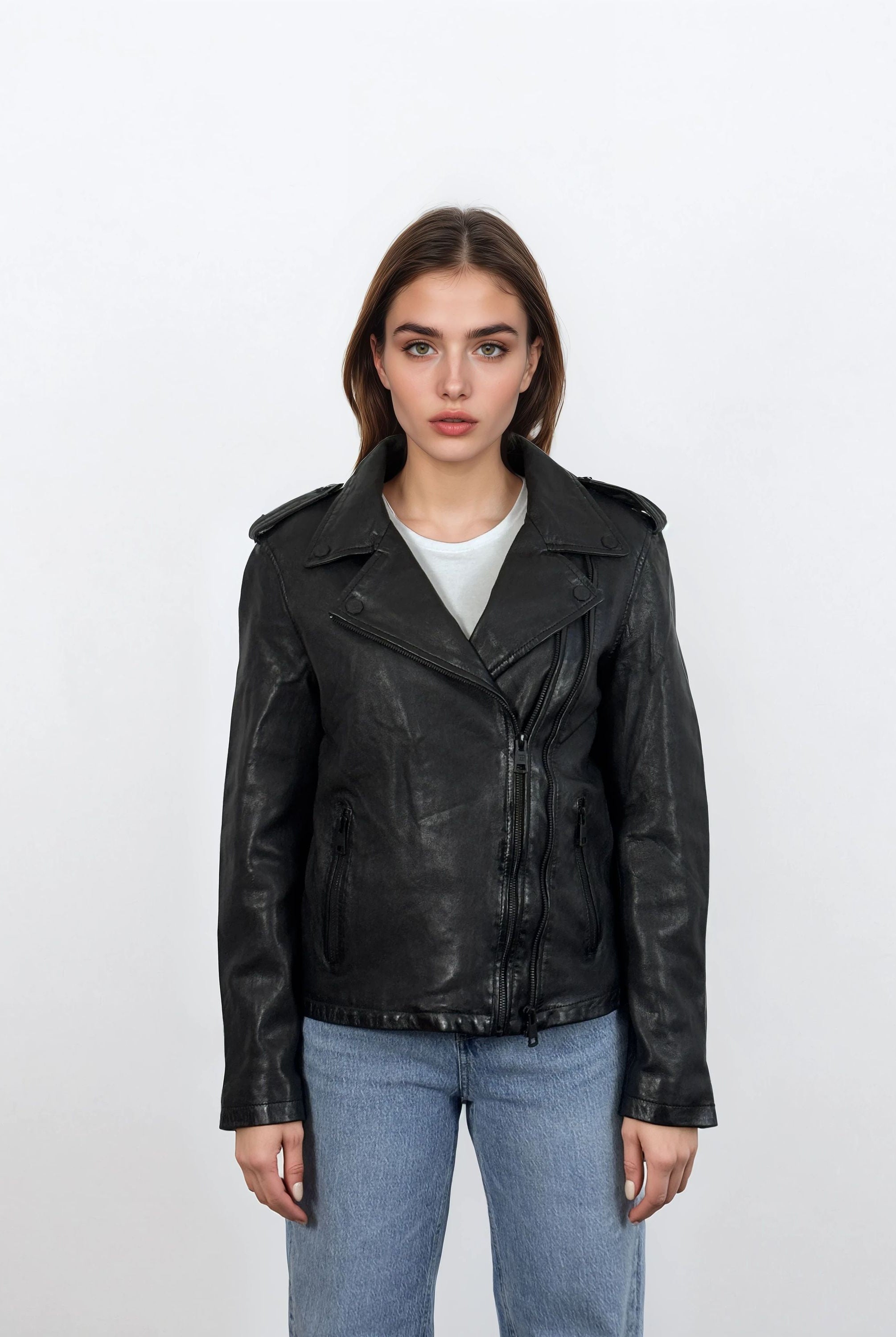 Eine Person steht mit dem Gesicht nach vorne vor einem weißen Hintergrund und trägt eine trueprodigy Damen Lederjacke aus Schafsleder in Schwarz im klassischen Biker-Stil über einem hellgrauen T-Shirt und blauen Jeans.