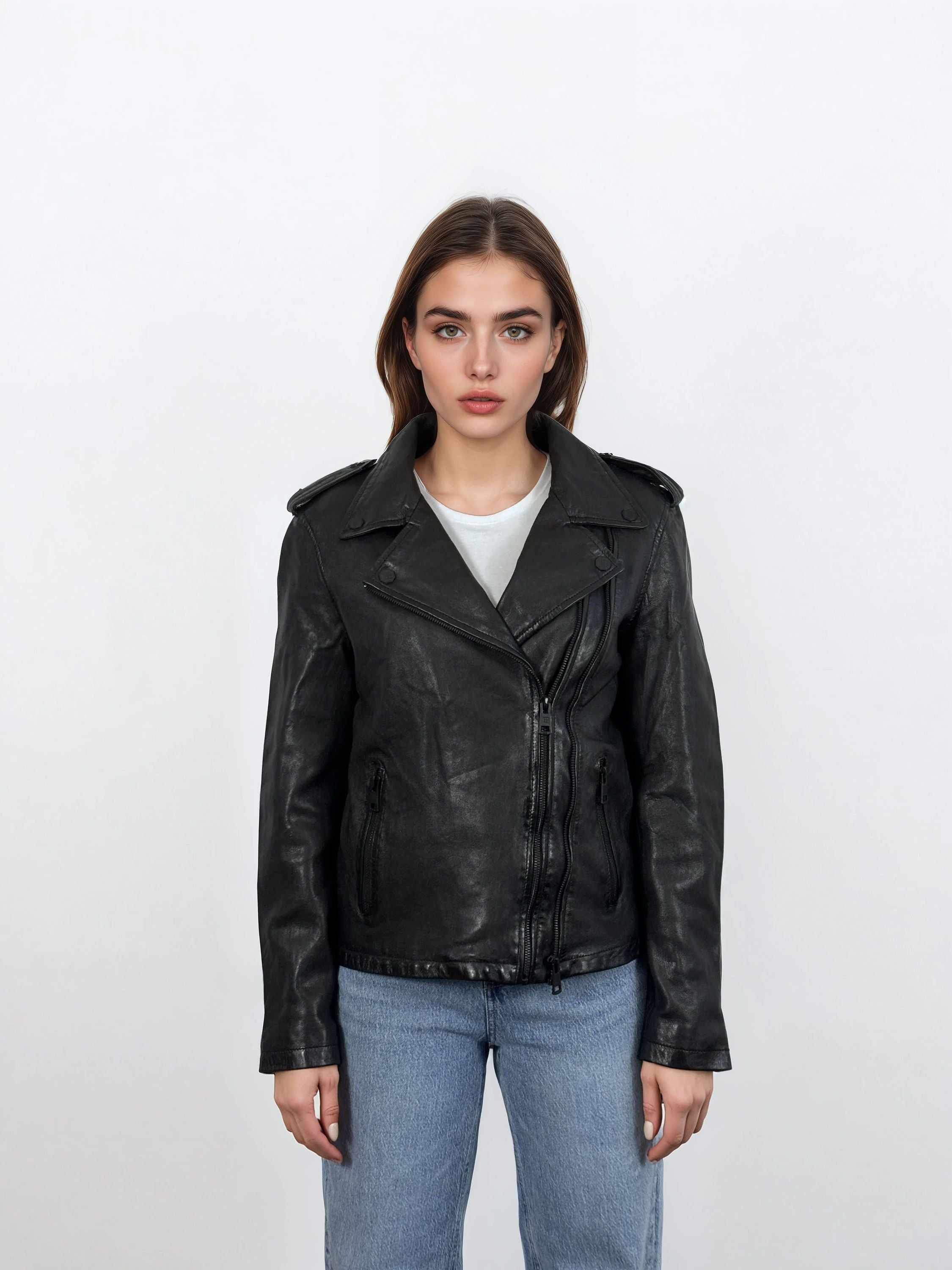 Eine Person steht mit dem Gesicht nach vorne vor einem weißen Hintergrund und trägt eine trueprodigy Damen Lederjacke aus Schafsleder in Schwarz im klassischen Biker-Stil über einem hellgrauen T-Shirt und blauen Jeans.