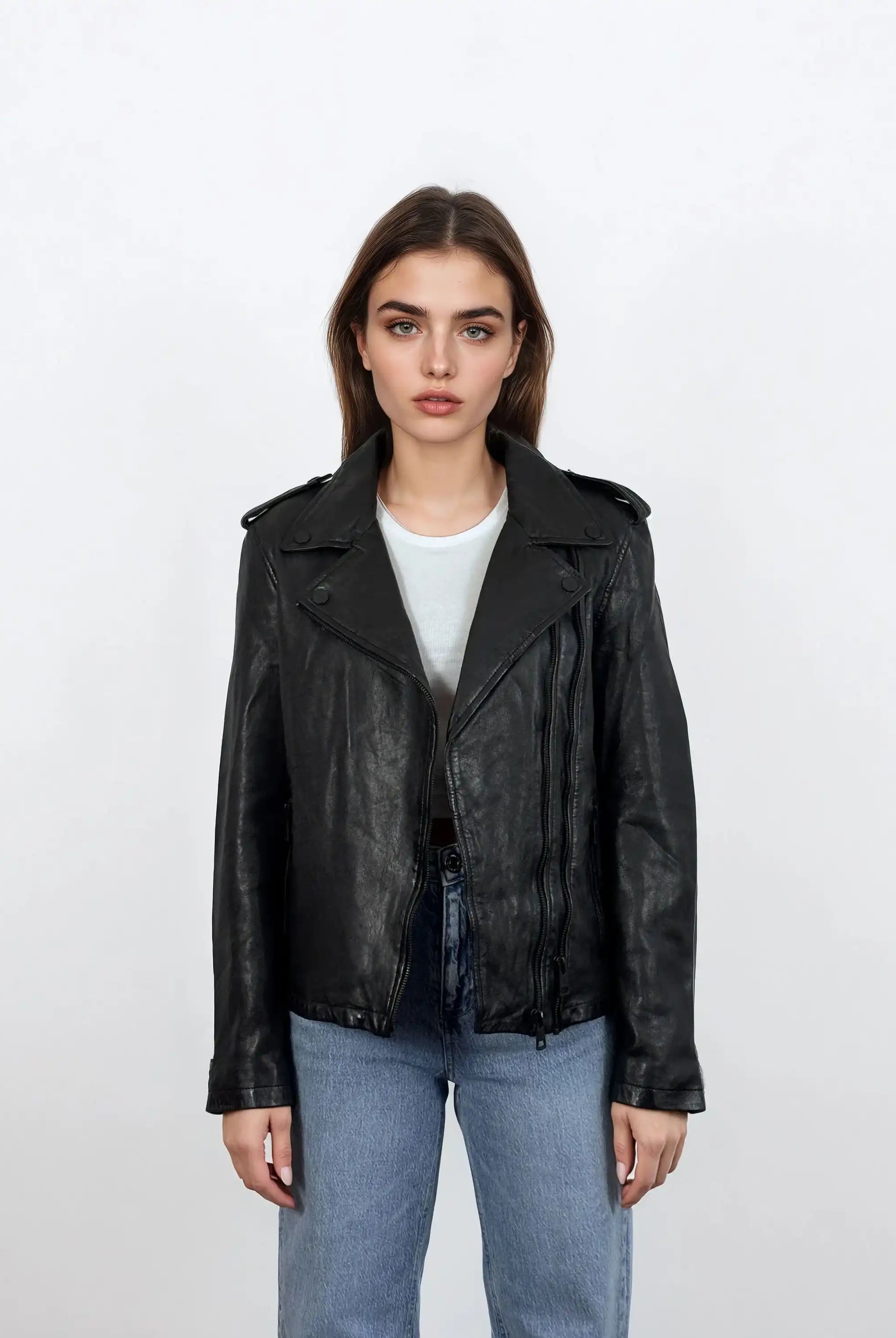 Eine Frau mit glattem braunem Haar trägt eine trueprodigy Damen Lederjacke aus Schafsleder in Schwarz im klassischen Biker-Stil über einem weißen T-Shirt und blauen Jeans und steht mit neutralem Gesichtsausdruck vor einem schlichten weißen Hintergrund.