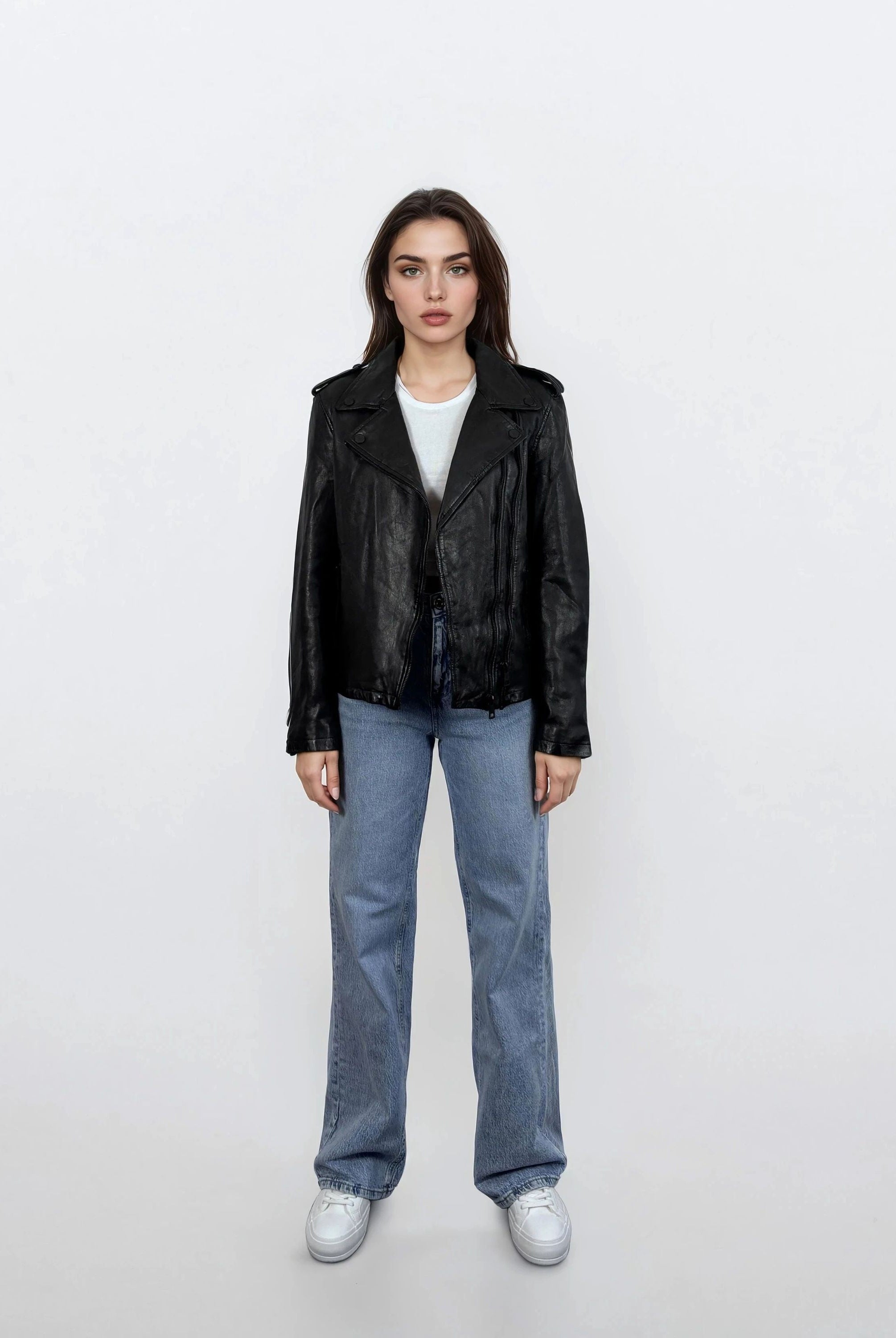 Eine Frau steht mit dem Gesicht nach vorne vor einem weißen Hintergrund und trägt eine trueprodigy Damen Lederjacke aus Schafsleder in Schwarz im klassischen Biker-Stil, gepaart mit einem weißen T-Shirt, blauen Jeans und weißen Sneakers. Ihre Arme sind an den Seiten verschränkt und ihr Gesichtsausdruck ist neutral.
