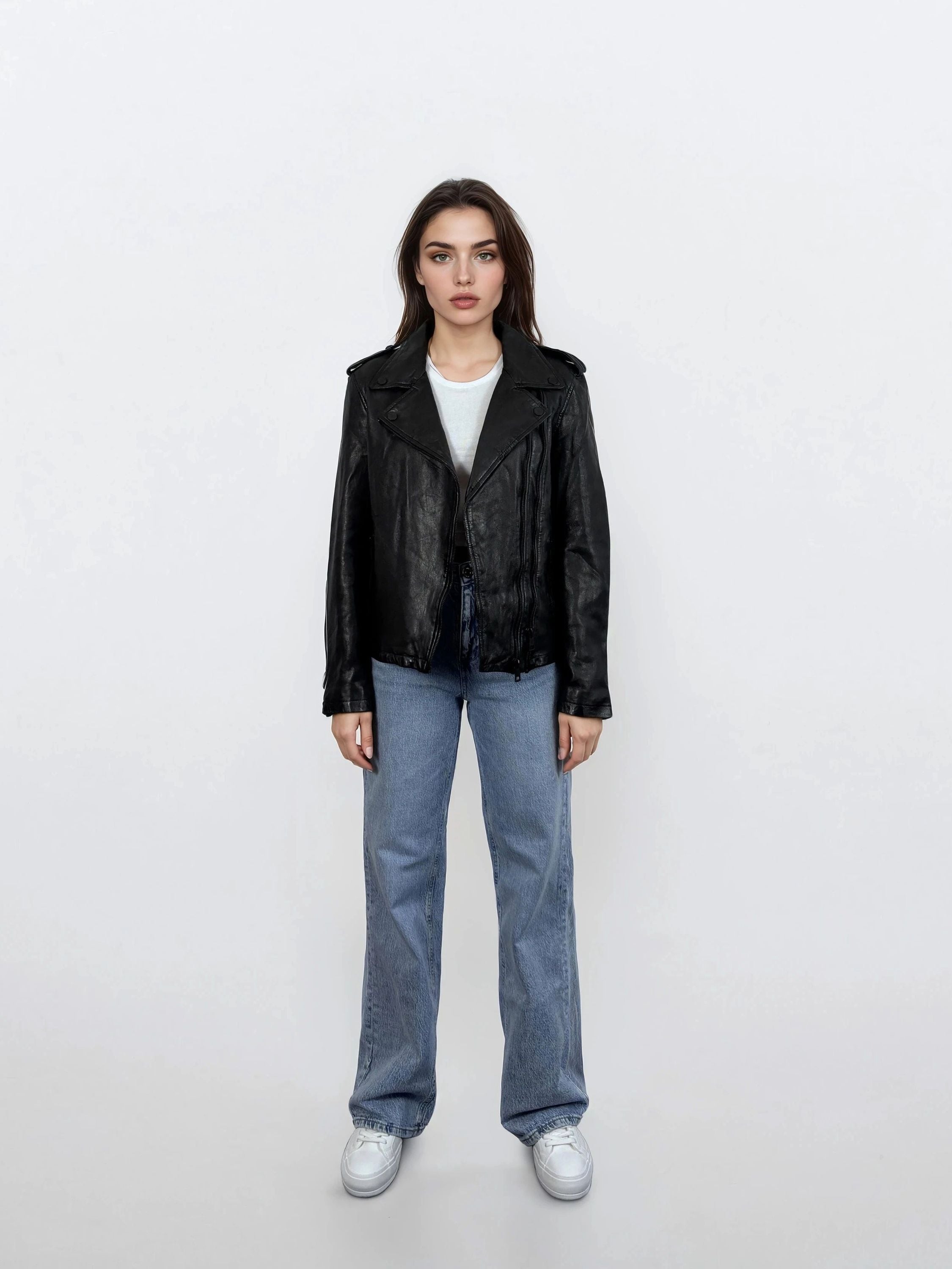 Eine Frau steht mit dem Gesicht nach vorne vor einem weißen Hintergrund und trägt eine trueprodigy Damen Lederjacke aus Schafsleder in Schwarz im klassischen Biker-Stil, gepaart mit einem weißen T-Shirt, blauen Jeans und weißen Sneakers. Ihre Arme sind an den Seiten verschränkt und ihr Gesichtsausdruck ist neutral.