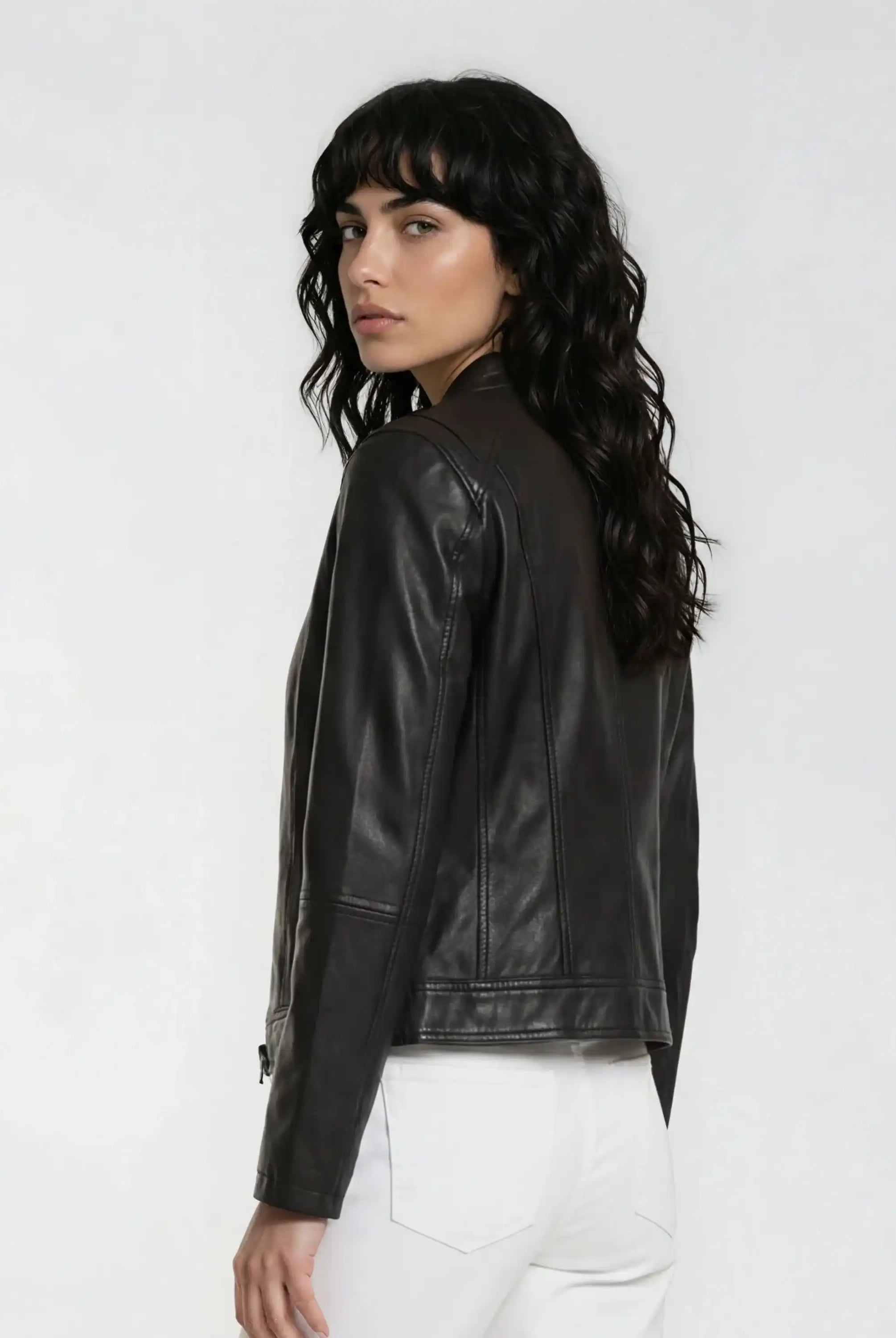 Eine Frau mit langen, gewellten dunklen Haaren trägt die trueprodigy Damen Lederjacke aus Schafsleder in Schwarz im modernen Café-Racer-Stil und eine weiße Hose, steht teilweise von der Kamera abgewandt vor einem hellen Hintergrund, der ihre weibliche Silhouette betont.