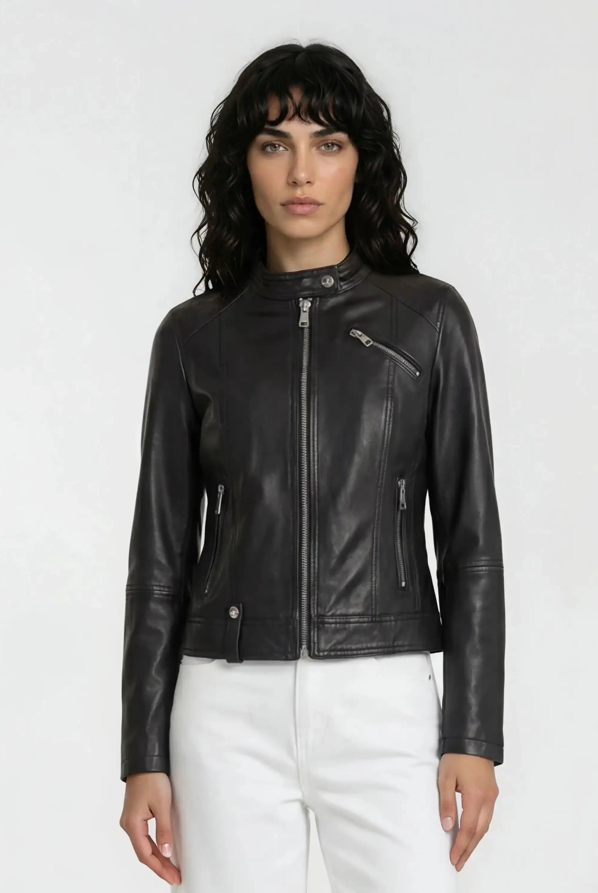 Eine Person mit langen lockigen dunklen Haaren trägt eine trueprodigy Damen Lederjacke aus Schafsleder in Schwarz im modernen Café-Racer-Stil mit Frontreißverschluss und Reißverschlusstaschen, gepaart mit einer weißen Hose, vor einem schlichten hellen Hintergrund.