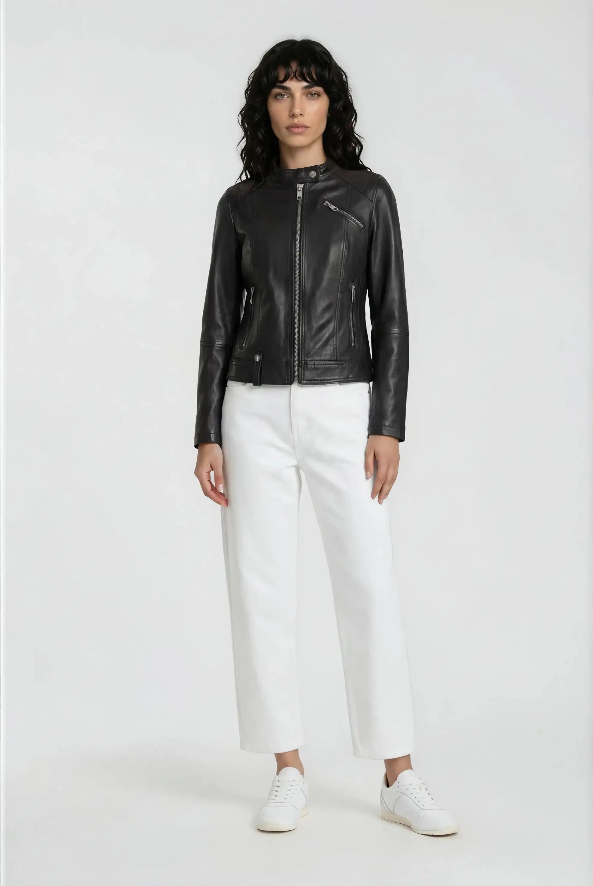 Eine Person steht vor einem schlichten Hintergrund und trägt die trueprodigy Damen Lederjacke aus Schafsleder in Schwarz im modernen Café-Racer-Stil, gepaart mit einer weißen, weit geschnittenen Hose und Sneakers. Sie hat dunkles, gewelltes Haar und eine moderne, feminine Silhouette.