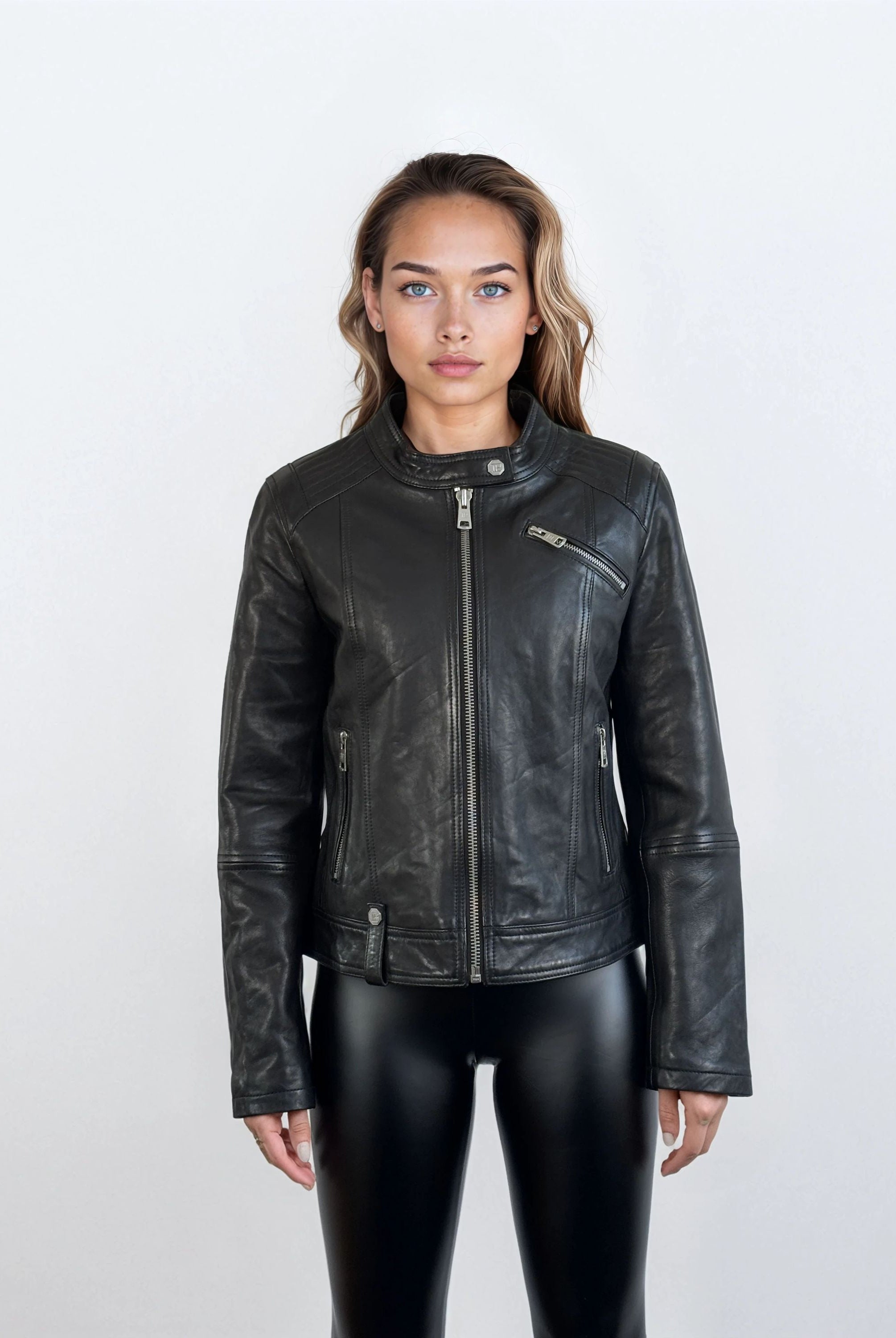 Eine Person steht mit dem Gesicht nach vorne vor einem schlichten, hellen Hintergrund und trägt die trueprodigy Damen Lederjacke aus Schafsleder in Schwarz im modernen Café-Racer-Stil mit Reißverschlusstaschen und schwarzer, glänzender Hose, die eine feminine Silhouette hervorhebt.