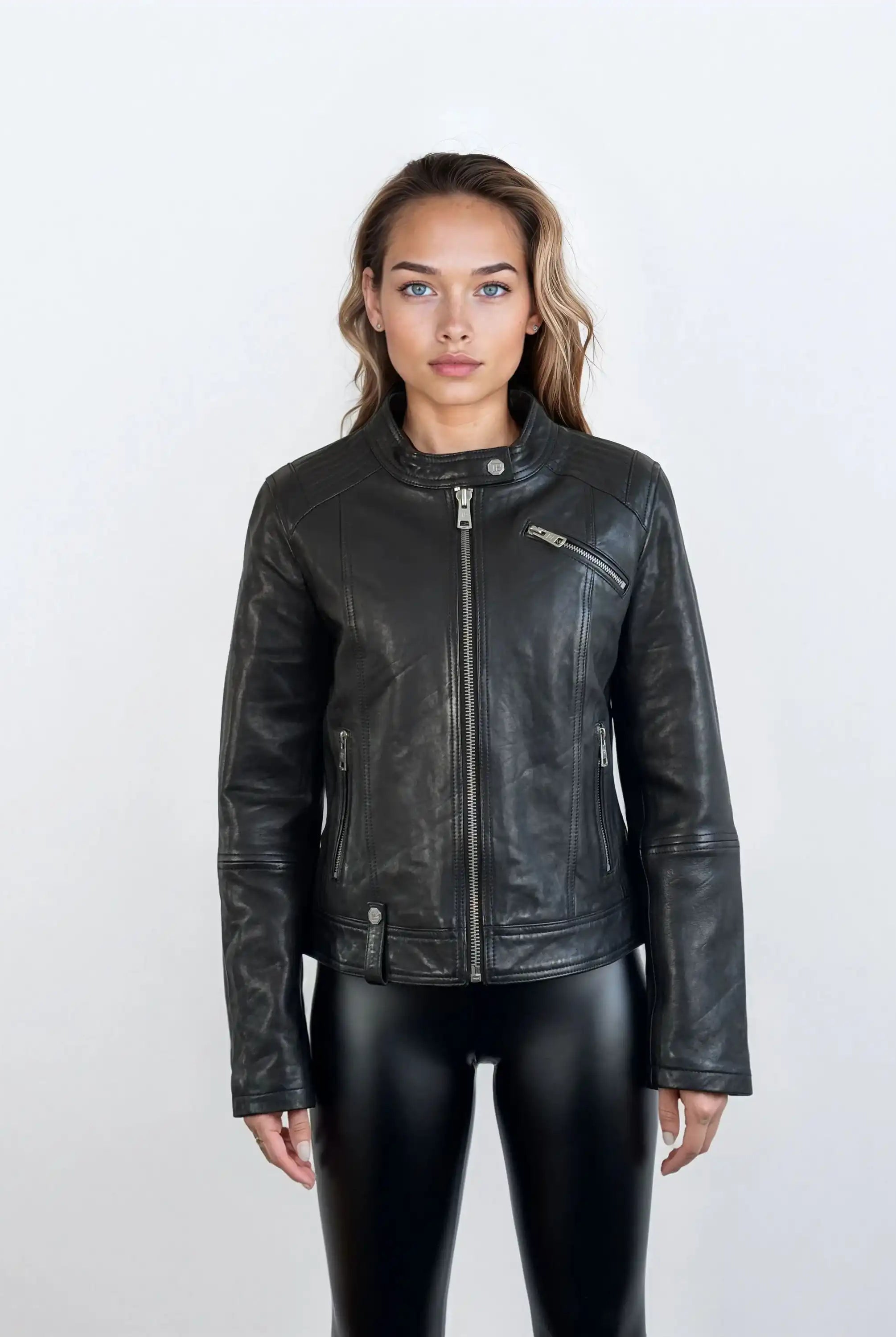 Eine Person steht mit dem Gesicht nach vorne vor einem schlichten, hellen Hintergrund und trägt die trueprodigy Damen Lederjacke aus Schafsleder in Schwarz im modernen Café-Racer-Stil mit Reißverschlusstaschen und schwarzer, glänzender Hose, die eine feminine Silhouette hervorhebt.