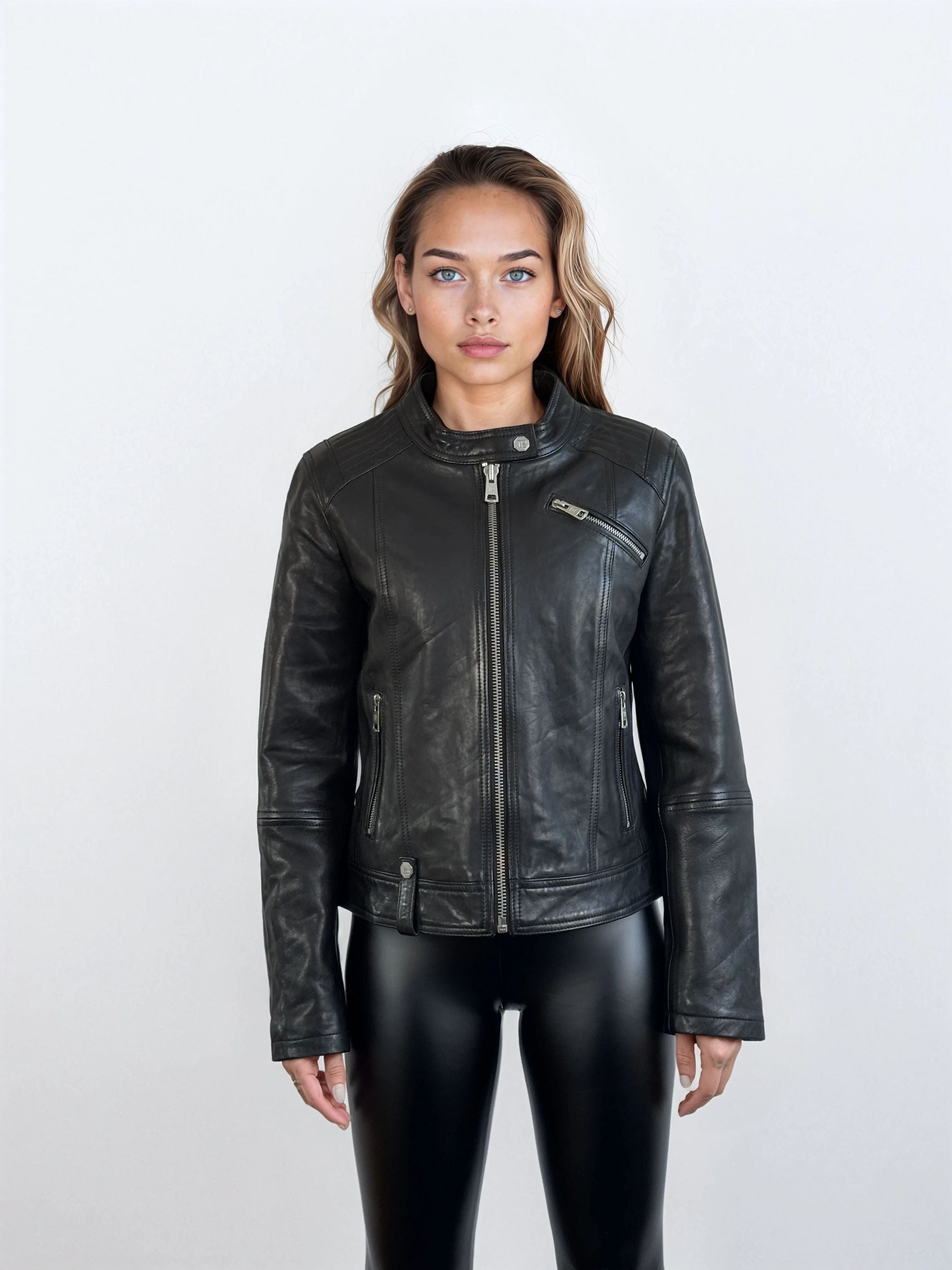Eine Person steht mit dem Gesicht nach vorne vor einem schlichten, hellen Hintergrund und trägt die trueprodigy Damen Lederjacke aus Schafsleder in Schwarz im modernen Café-Racer-Stil mit Reißverschlusstaschen und schwarzer, glänzender Hose, die eine feminine Silhouette hervorhebt.