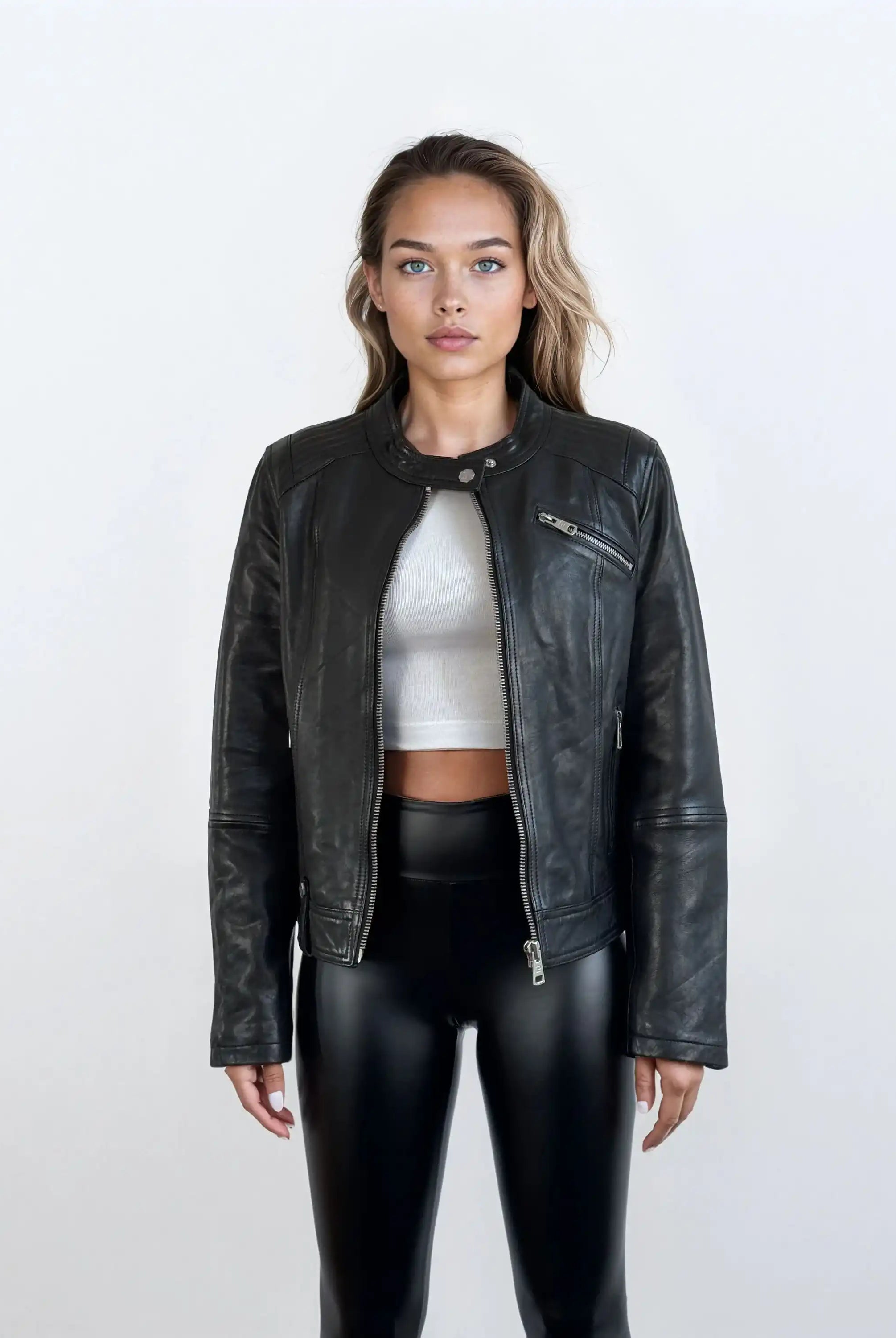 Eine Frau trägt die schwarze Damen Lederjacke aus Schafsleder im modernen Café-Racer-Stil von trueprodigy über einem weißen Crop-Top und glänzenden schwarzen Hosen vor einem schlichten hellen Hintergrund.