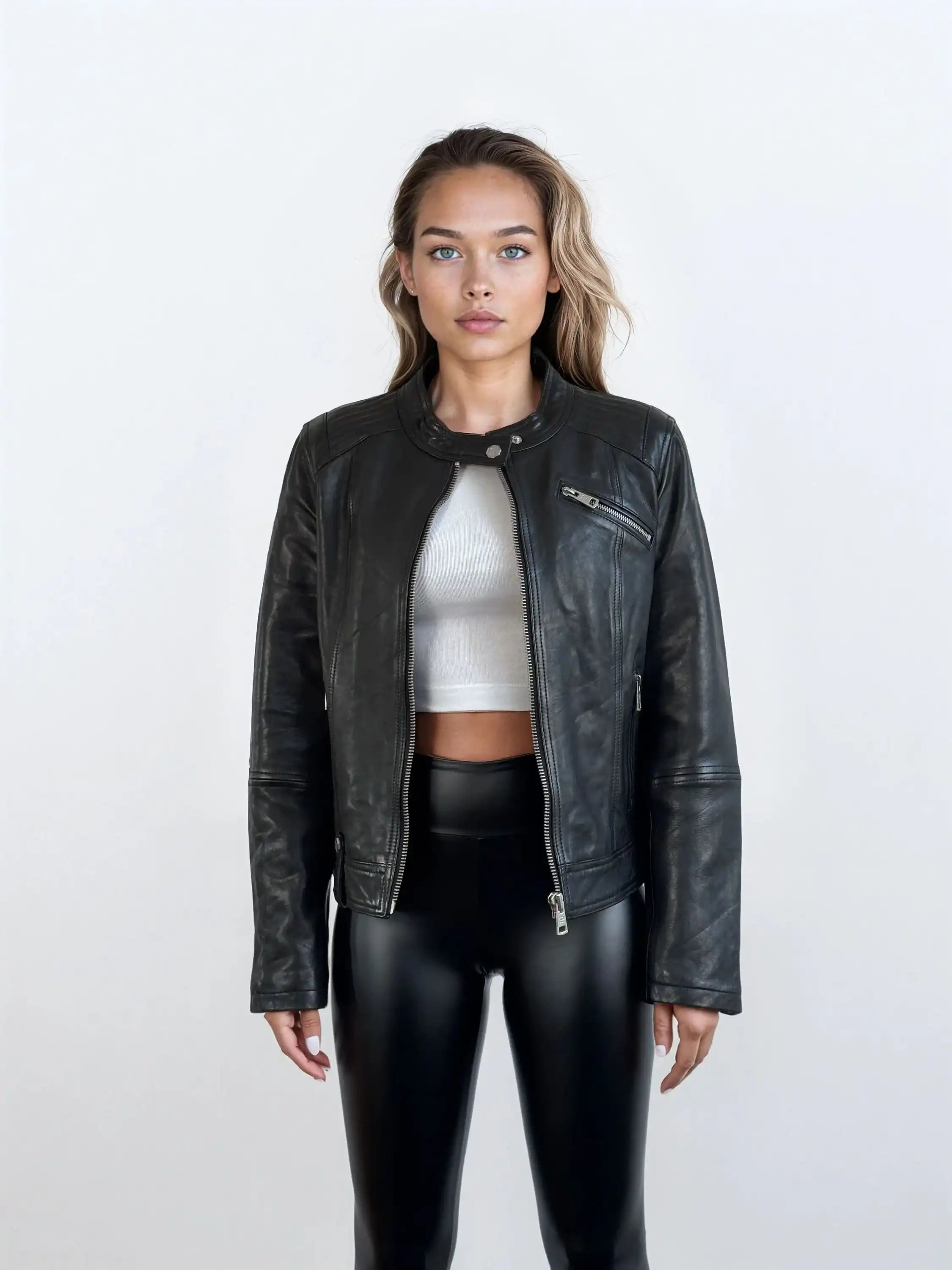 Eine Frau trägt die schwarze Damen Lederjacke aus Schafsleder im modernen Café-Racer-Stil von trueprodigy über einem weißen Crop-Top und glänzenden schwarzen Hosen vor einem schlichten hellen Hintergrund.