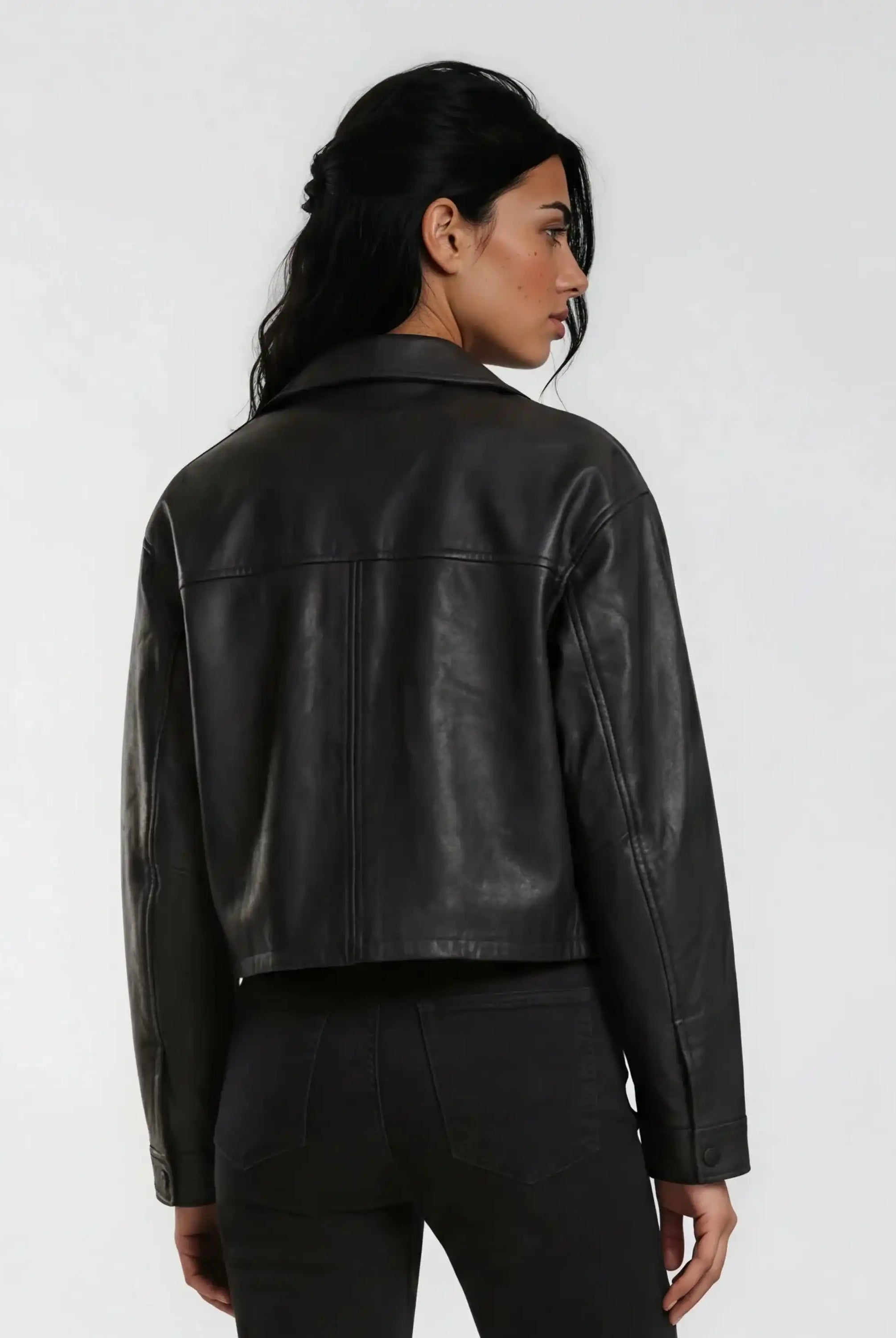 Eine Person mit langen dunklen Haaren trägt die trueprodigy Damen Lederjacke aus Schafsleder in Schwarz im Oversized Cropped-Style und schwarze Jeans und steht mit dem Rücken zur Kamera vor einem schlichten hellen Hintergrund.