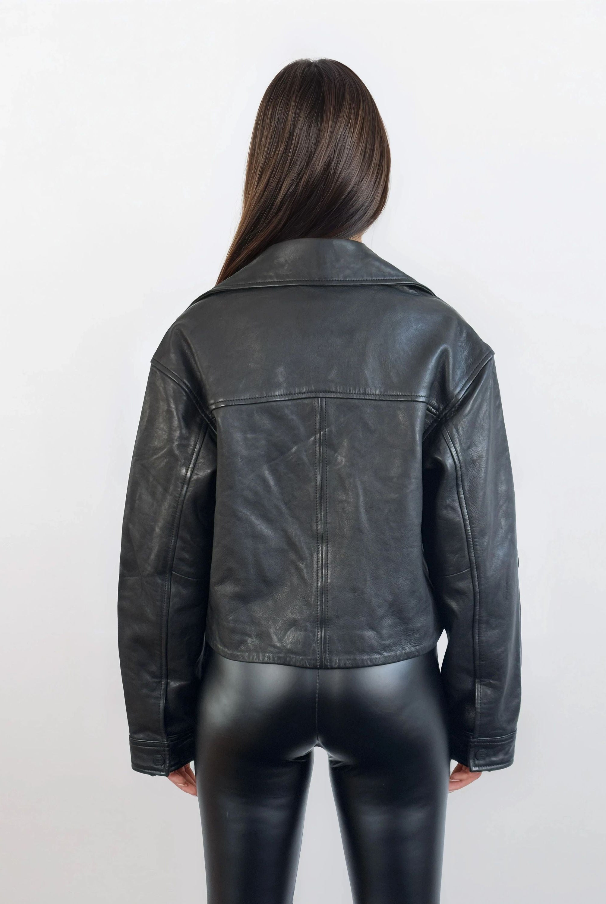 Eine Frau mit langen braunen Haaren steht mit der trueprodigy Damen Lederjacke aus Schafsleder in Schwarz im Oversized Cropped-Style und einer glänzenden schwarzen Hose vor einem schlichten hellen Hintergrund.