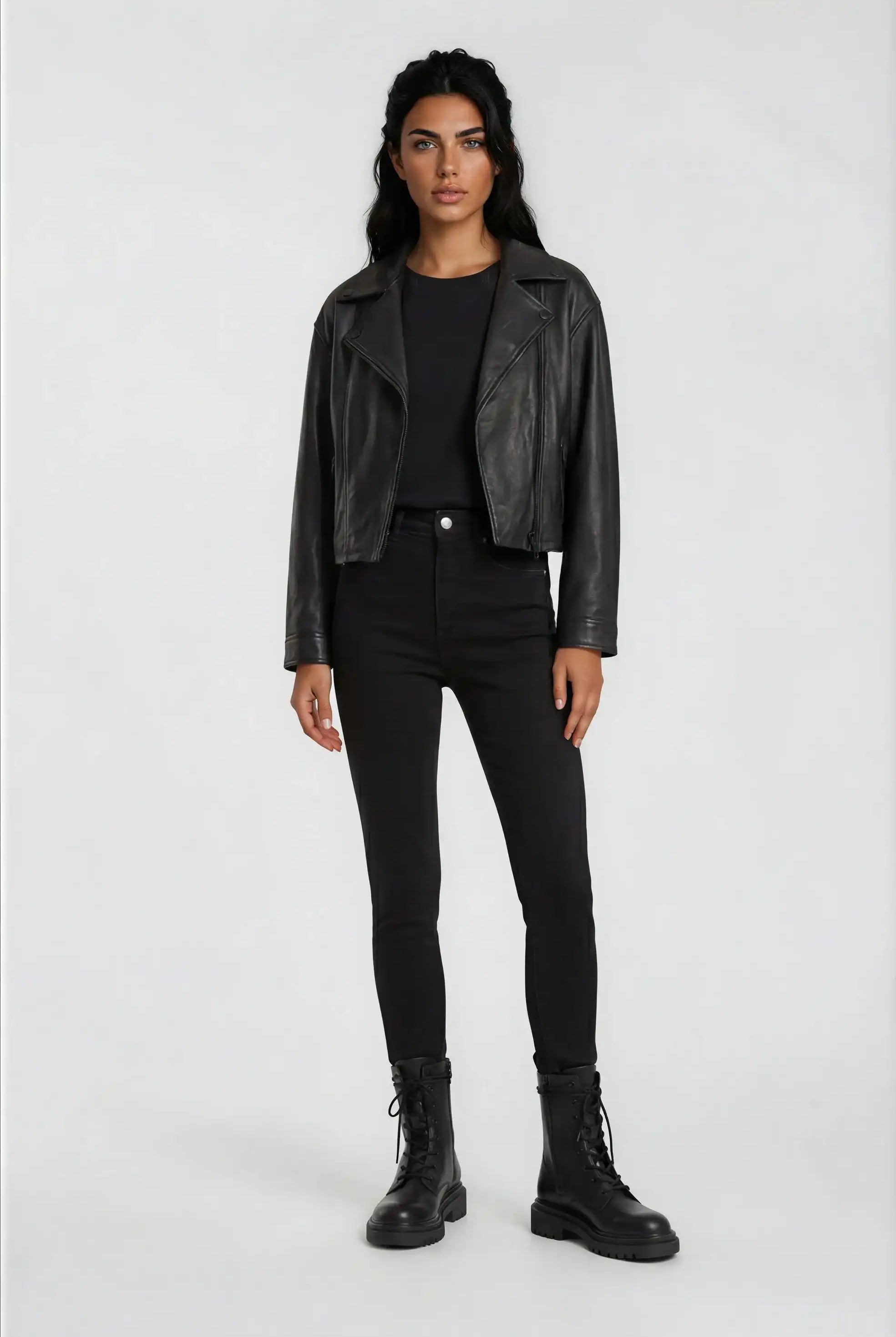 Eine Person steht mit dem Gesicht nach vorne vor einem unifarbenen Hintergrund und trägt eine trueprodigy Damen Lederjacke aus Schafsleder in Schwarz im Oversized Cropped-Style, ein schwarzes T-Shirt, High-Waisted Skinny Jeans und schwarze Schnürstiefel.