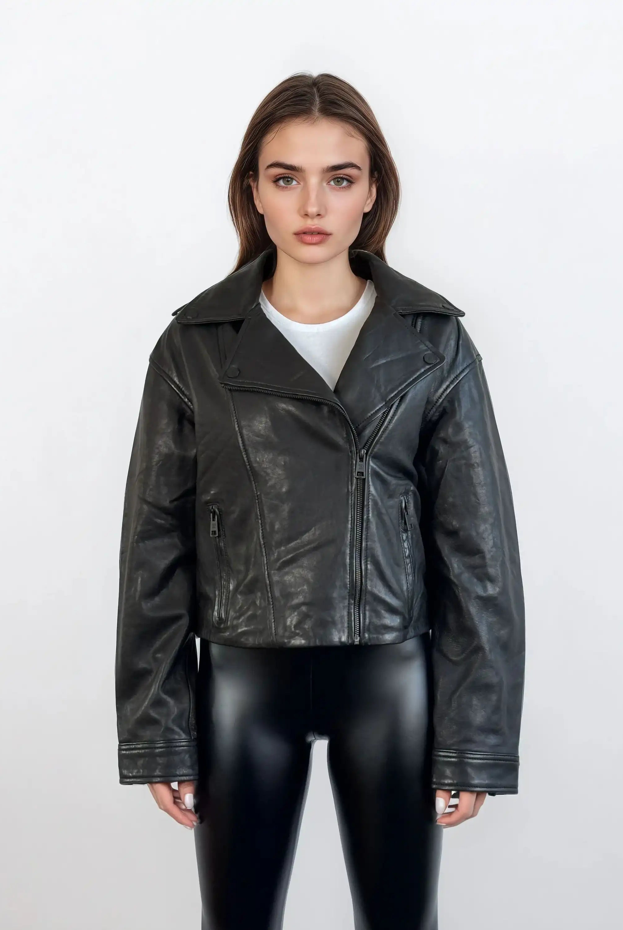 Eine Frau steht vor einem weißen Hintergrund und trägt die trueprodigy Damen Lederjacke aus Schafsleder in Schwarz im Oversized Cropped-Style über einer weißen Bluse und glänzenden schwarzen Leggings und schaut mit verschränkten Armen in die Kamera.