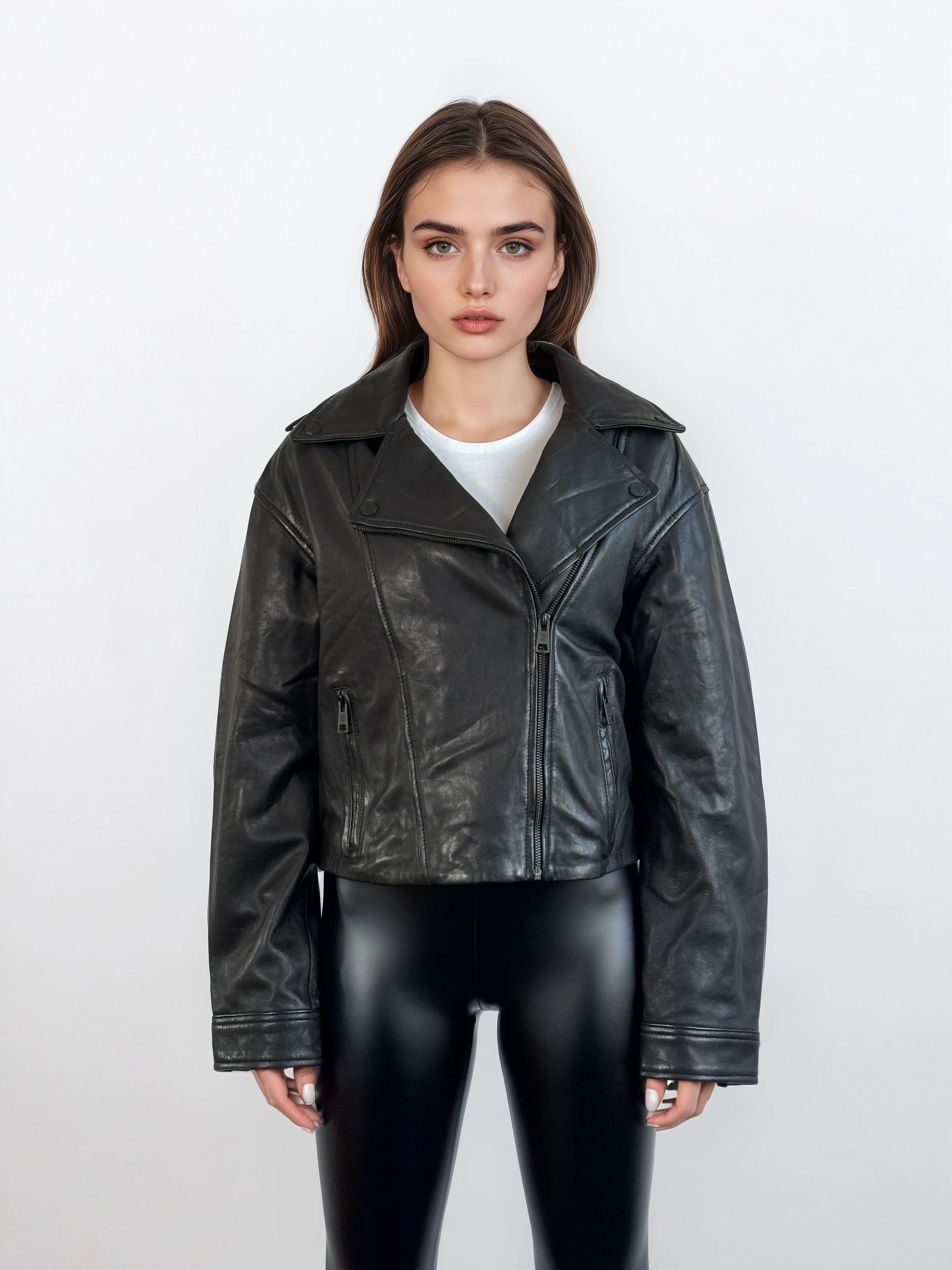 Eine Frau steht vor einem weißen Hintergrund und trägt die trueprodigy Damen Lederjacke aus Schafsleder in Schwarz im Oversized Cropped-Style über einer weißen Bluse und glänzenden schwarzen Leggings und schaut mit verschränkten Armen in die Kamera.