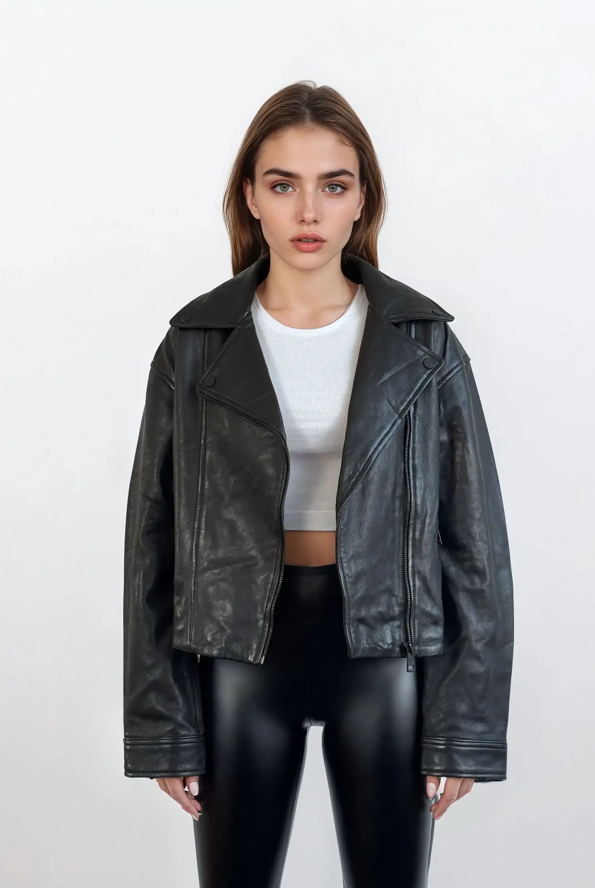 Eine Frau trägt die trueprodigy Damen Lederjacke aus Schafsleder in Schwarz im Oversized Cropped-Style über einem weißen Crop-Top und glänzenden schwarzen Hosen; sie steht vor weißem Hintergrund und blickt neutral nach vorn.
