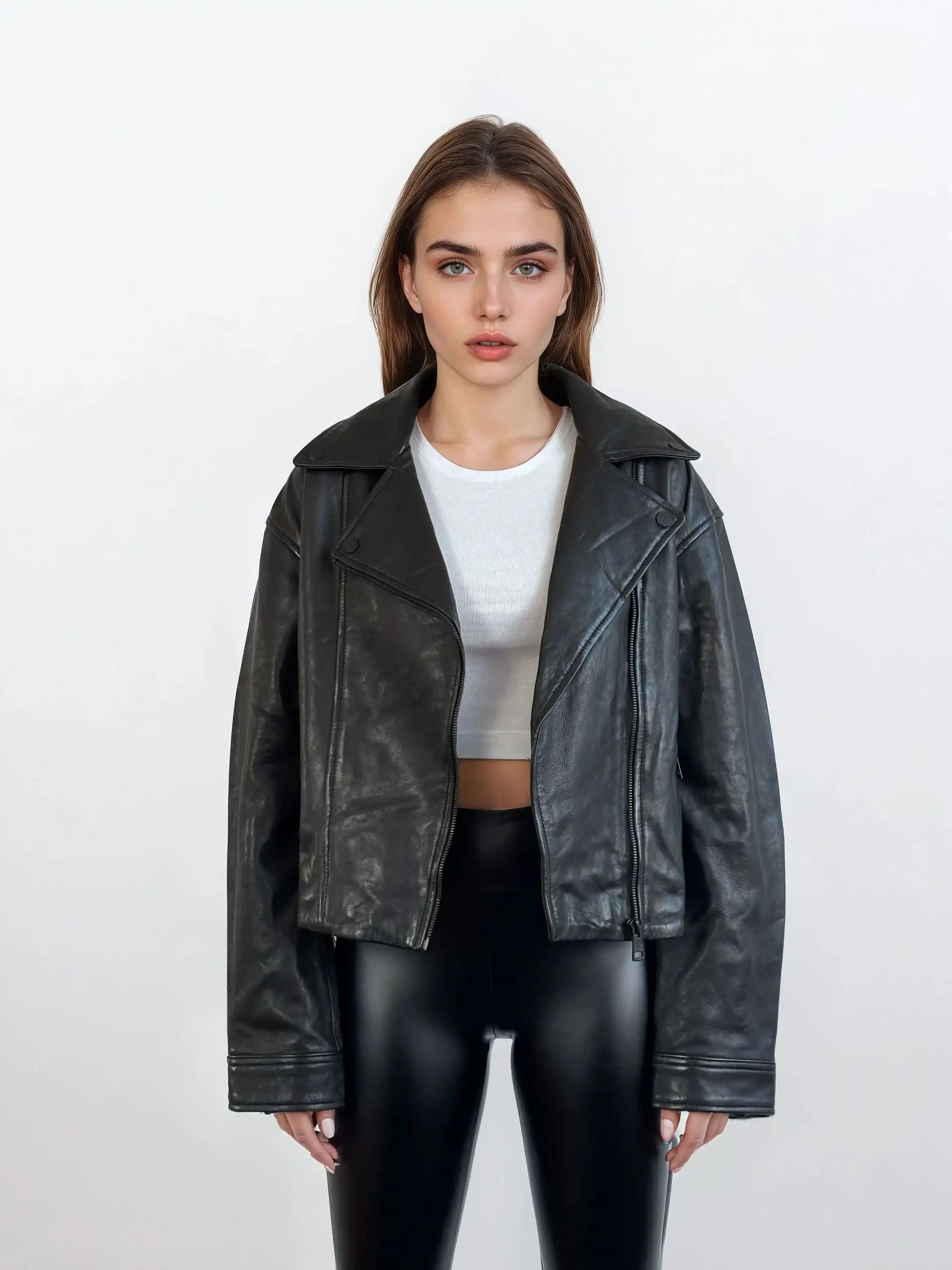 Eine Frau trägt die trueprodigy Damen Lederjacke aus Schafsleder in Schwarz im Oversized Cropped-Style über einem weißen Crop-Top und glänzenden schwarzen Hosen; sie steht vor weißem Hintergrund und blickt neutral nach vorn.