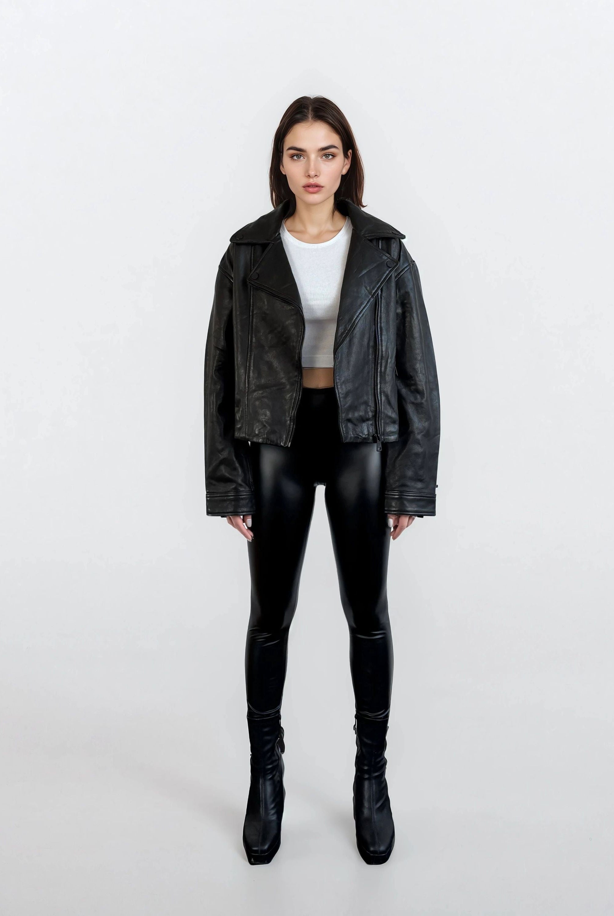 Eine Frau steht vor einem weißen Hintergrund und trägt die trueprodigy Damen Lederjacke aus Schafsleder in Schwarz im Oversized Cropped-Style, gepaart mit einem weißen Cropped-Top, schwarzen, glänzenden Leggings und Stiefeln mit Absatz und blickt neutral in die Kamera.