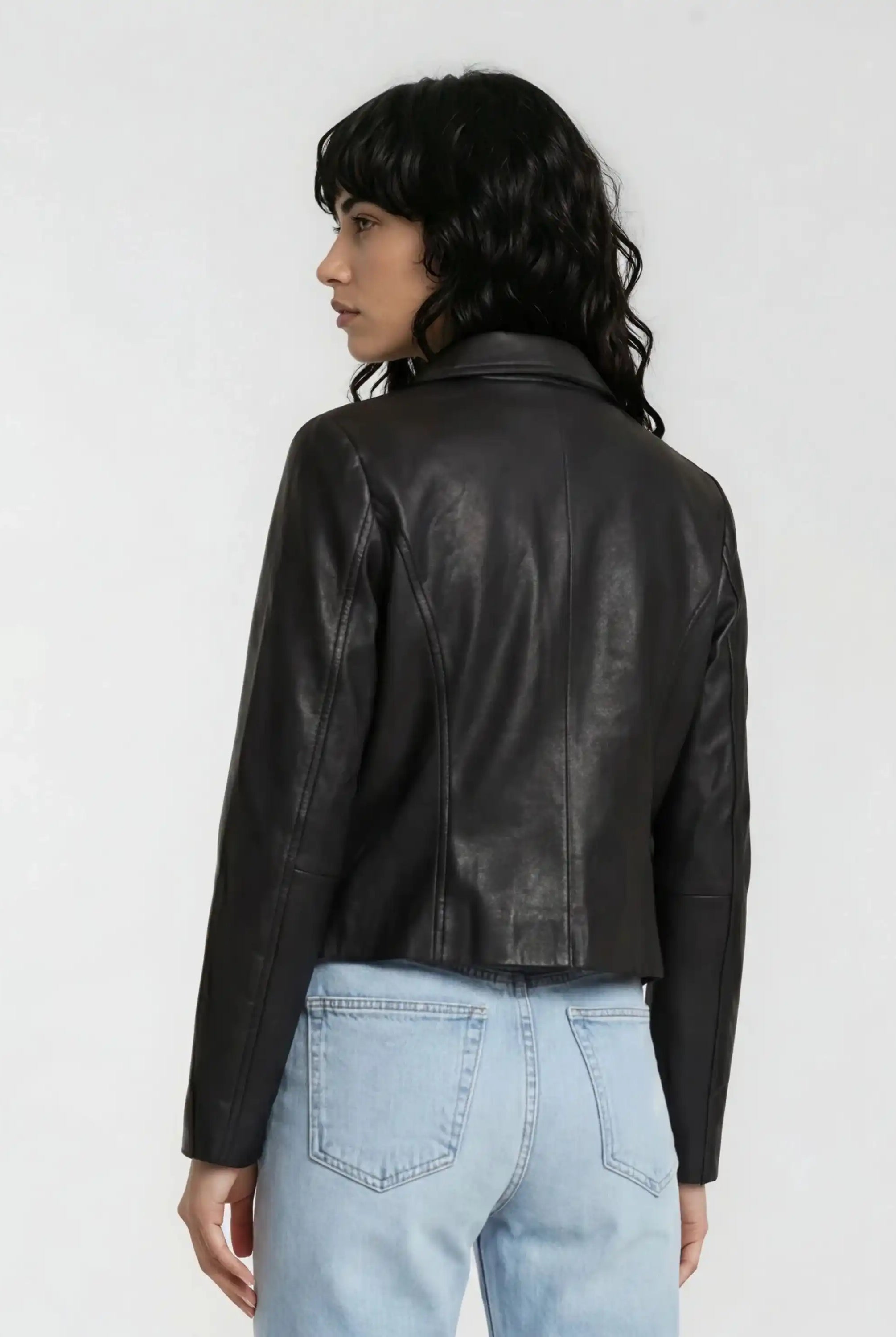 Eine Person mit langem, dunklem, welligem Haar trägt vor hellem Hintergrund eine schwarze trueprodigy Damen Lederjacke aus Schafsleder mit minimalistischem Design und hellblauen Jeans im Blouson-Stil.