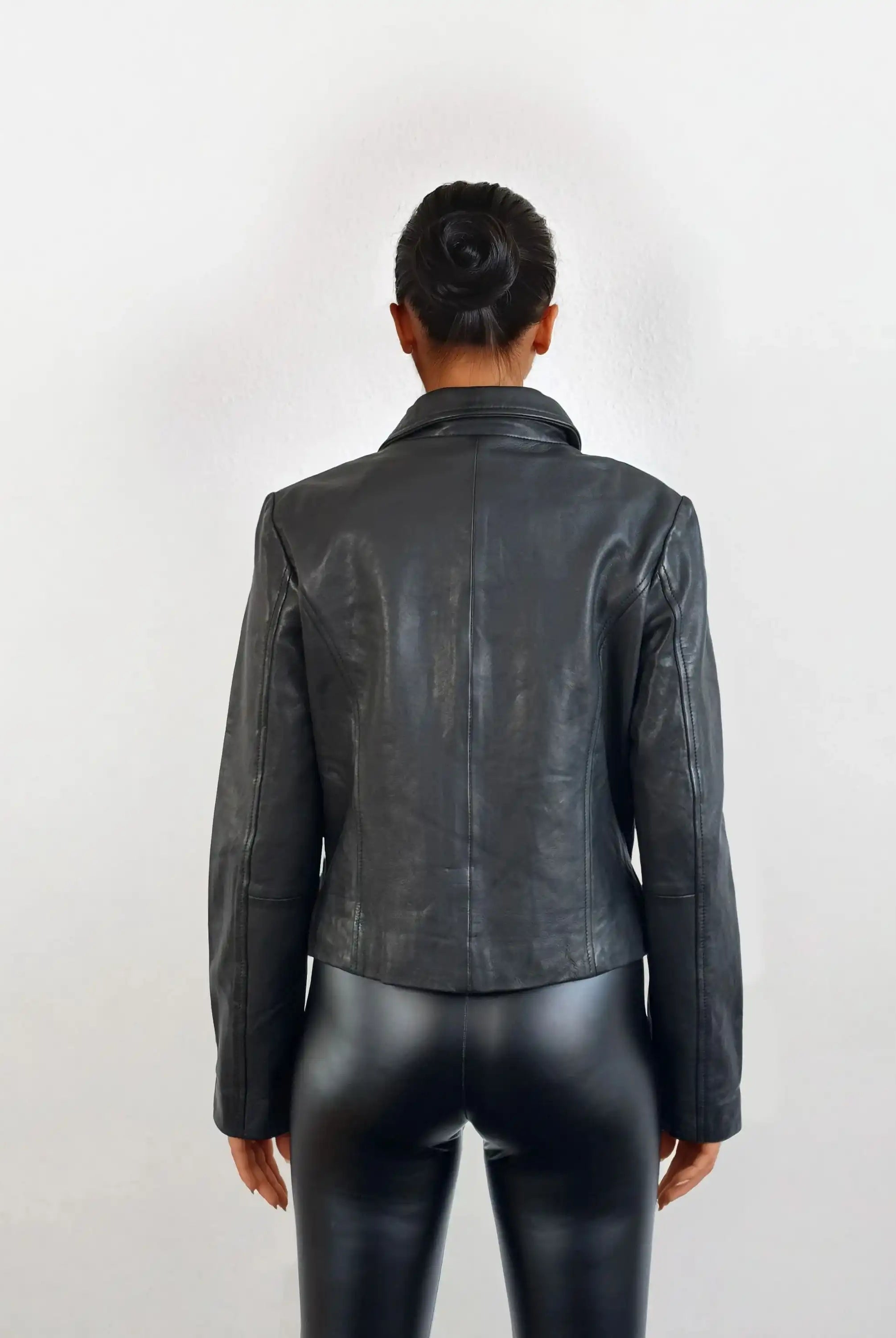 Eine Person mit ordentlich gebundenem Dutt steht vor weißem Hintergrund und trägt die schwarze Damen Lederjacke aus Schafsleder mit minimalistischem Design von trueprodigy sowie schwarze Lederhosen.