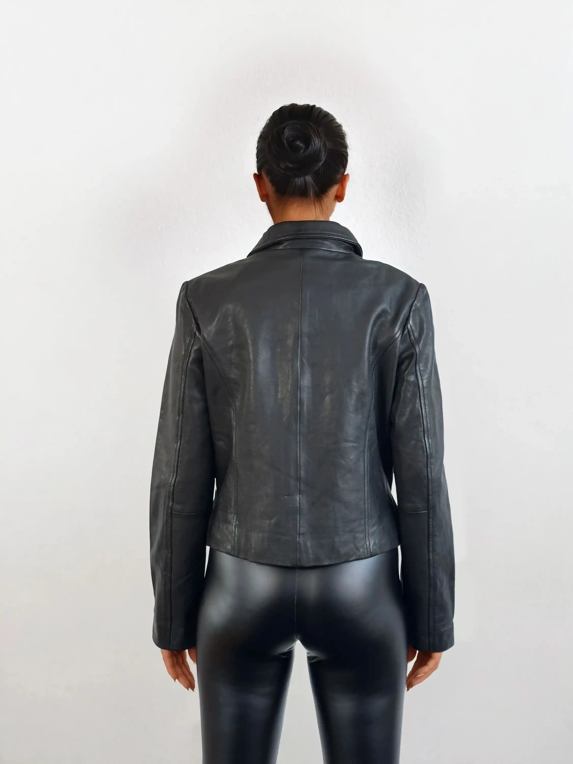 Eine Person mit ordentlich gebundenem Dutt steht vor weißem Hintergrund und trägt die schwarze Damen Lederjacke aus Schafsleder mit minimalistischem Design von trueprodigy sowie schwarze Lederhosen.