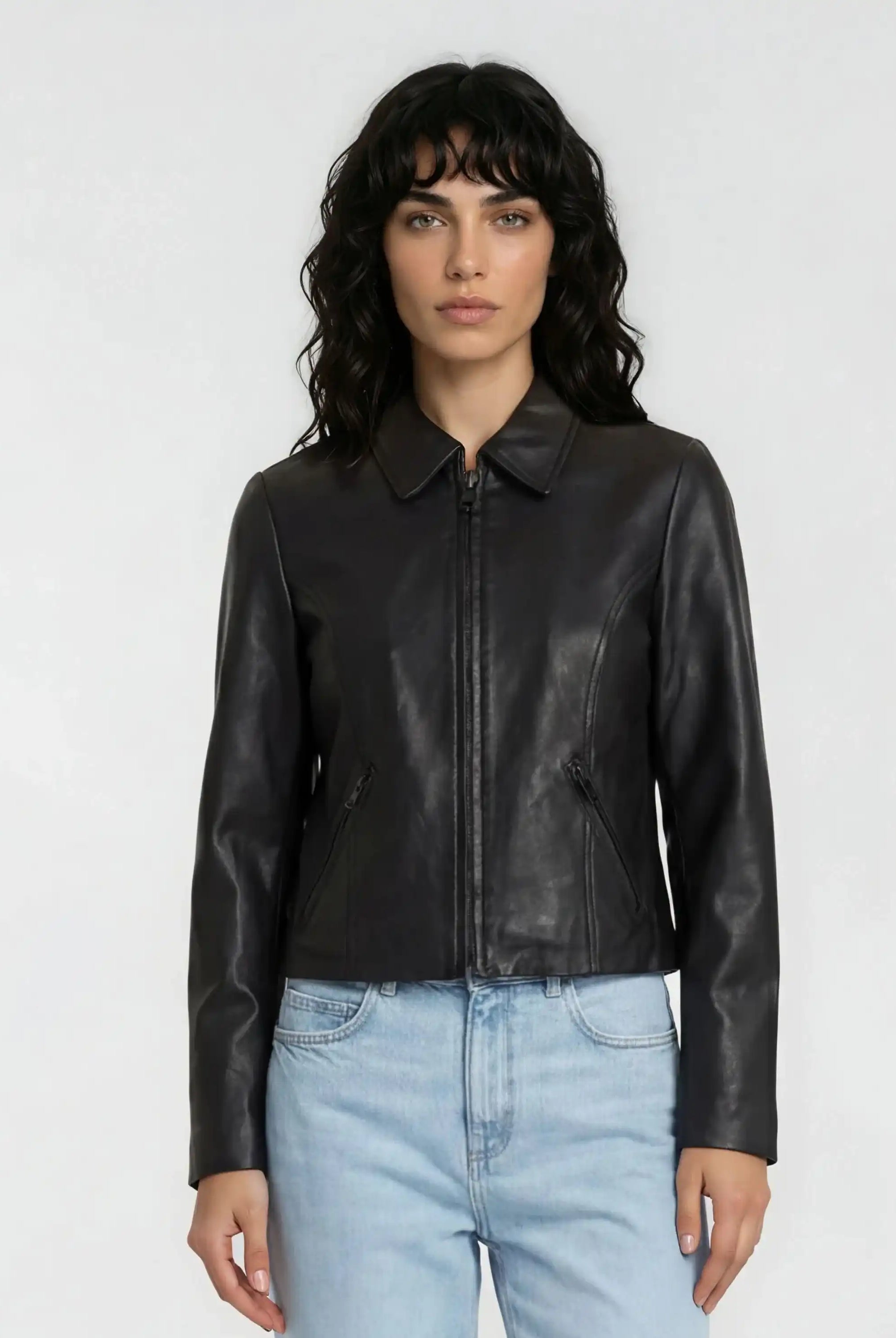 Eine Person mit gewelltem dunklem Haar trägt eine trueprodigy Damen Lederjacke aus Schafsleder in Schwarz mit minimalistischem Design und hellblaue Jeans vor einem schlichten hellen Hintergrund.