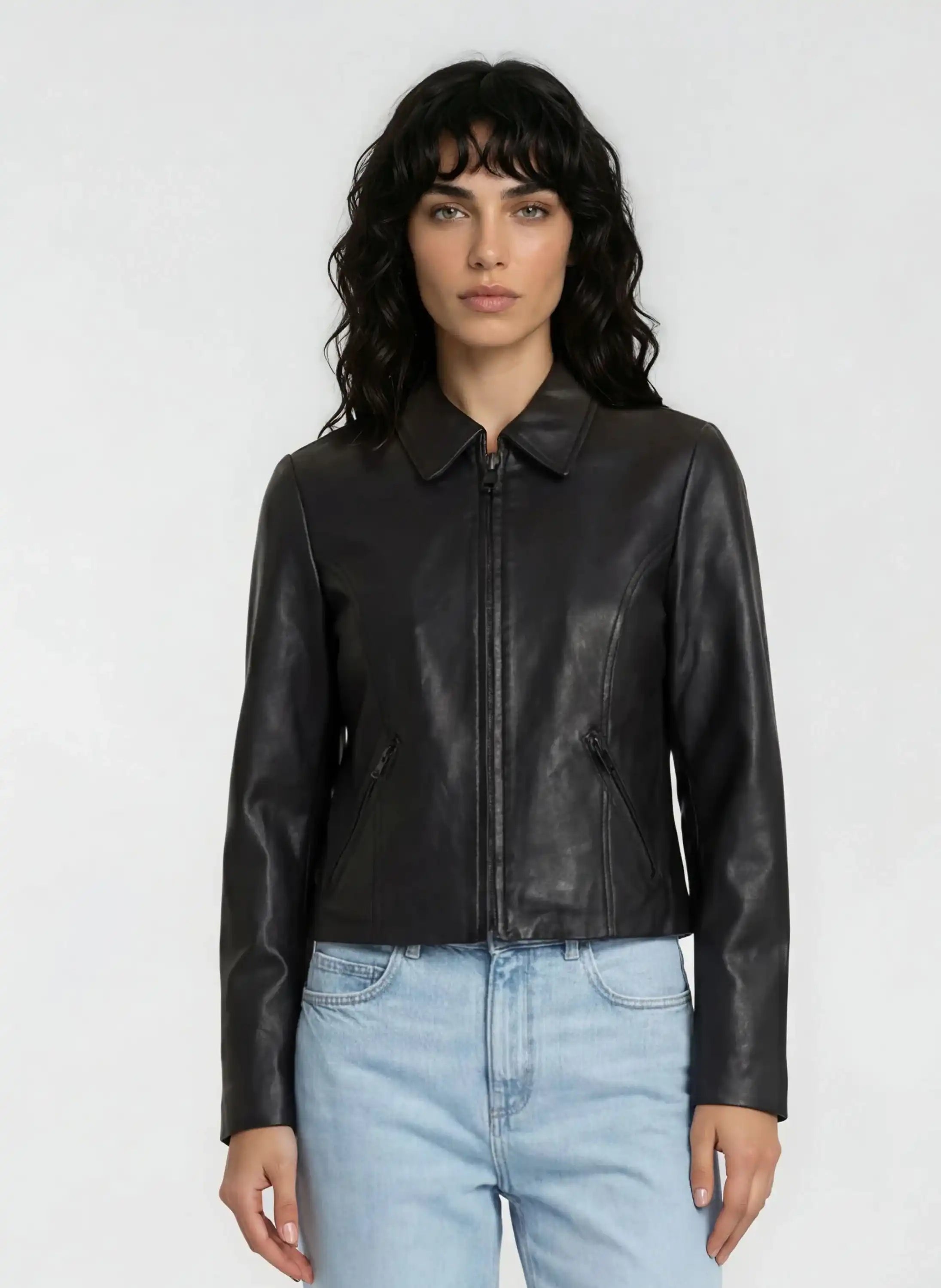 Eine Person mit gewelltem dunklem Haar trägt eine trueprodigy Damen Lederjacke aus Schafsleder in Schwarz mit minimalistischem Design und hellblaue Jeans vor einem schlichten hellen Hintergrund.