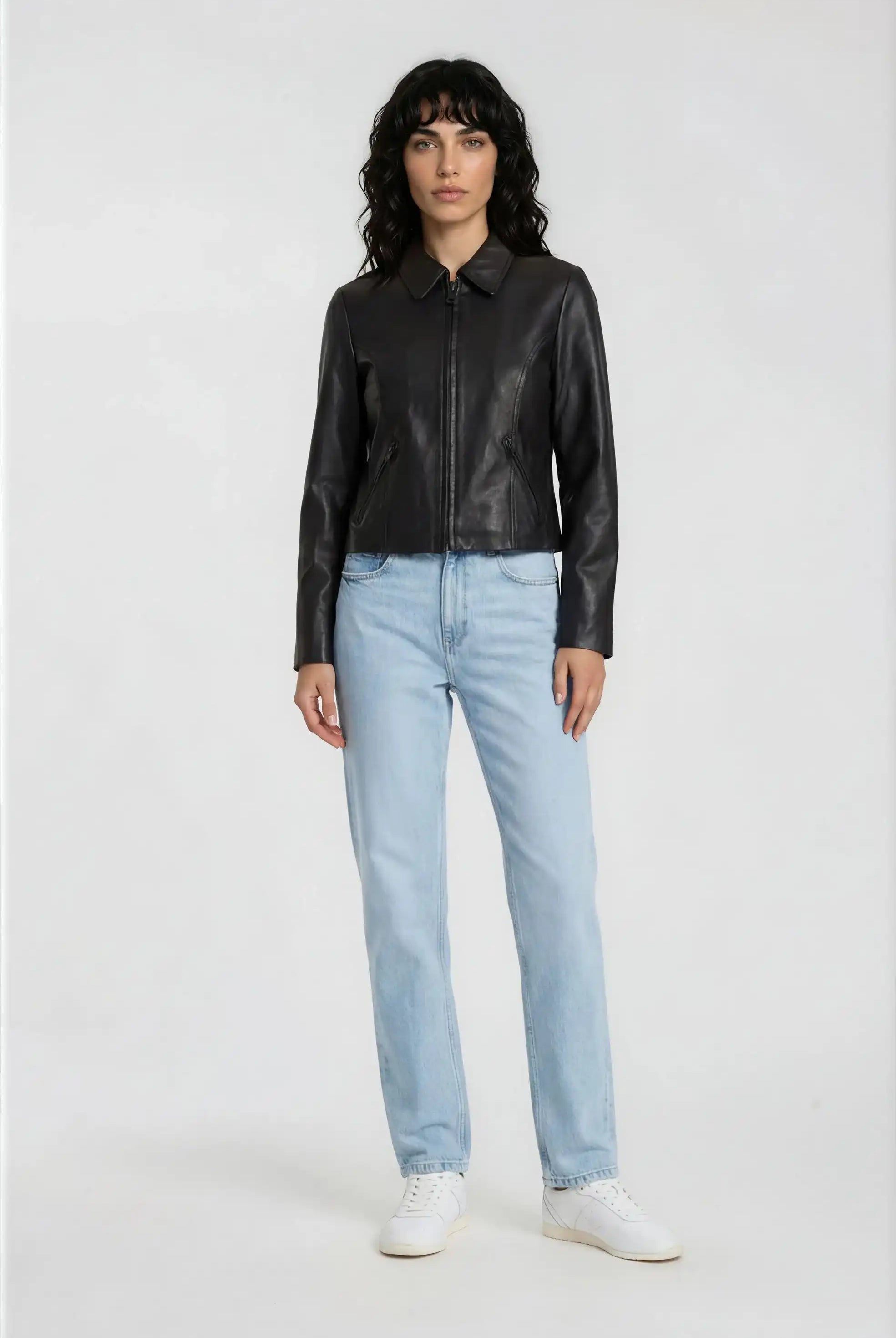 Eine Frau trägt die trueprodigy Damen Lederjacke aus Schafsleder in Schwarz mit minimalistischem Design, kombiniert mit hellblauen Jeans und weißen Sneakern, vor schlicht weißem Hintergrund.