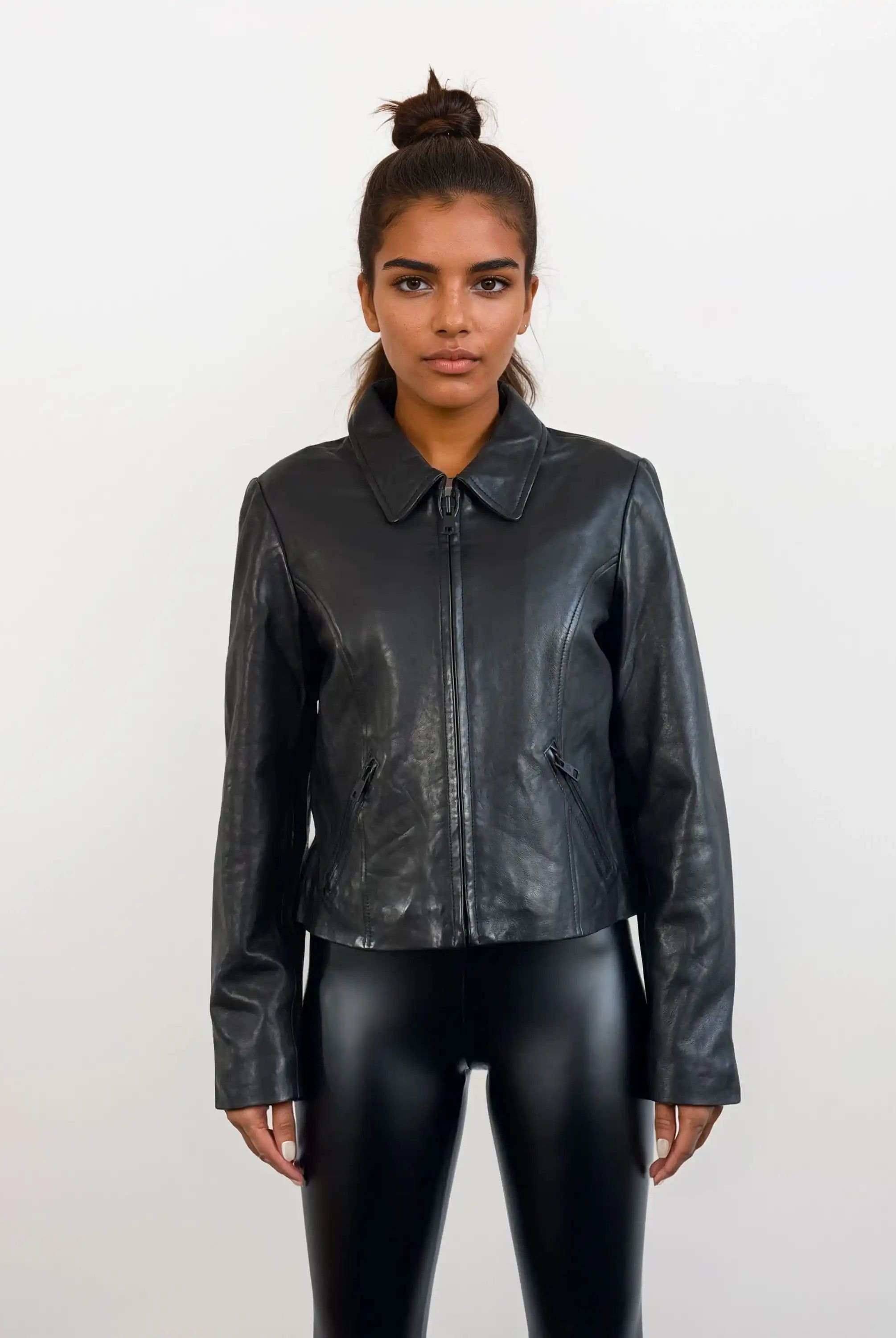 Eine Person steht mit dem Gesicht nach vorne vor einem weißen Hintergrund und trägt die trueprodigy Damen Lederjacke aus Schafsleder in Schwarz mit minimalistischem Design und schwarz glänzenden Leggings. Ihr Haar ist zu einem Dutt hochgesteckt, die Arme ruhen natürlich an der Seite.