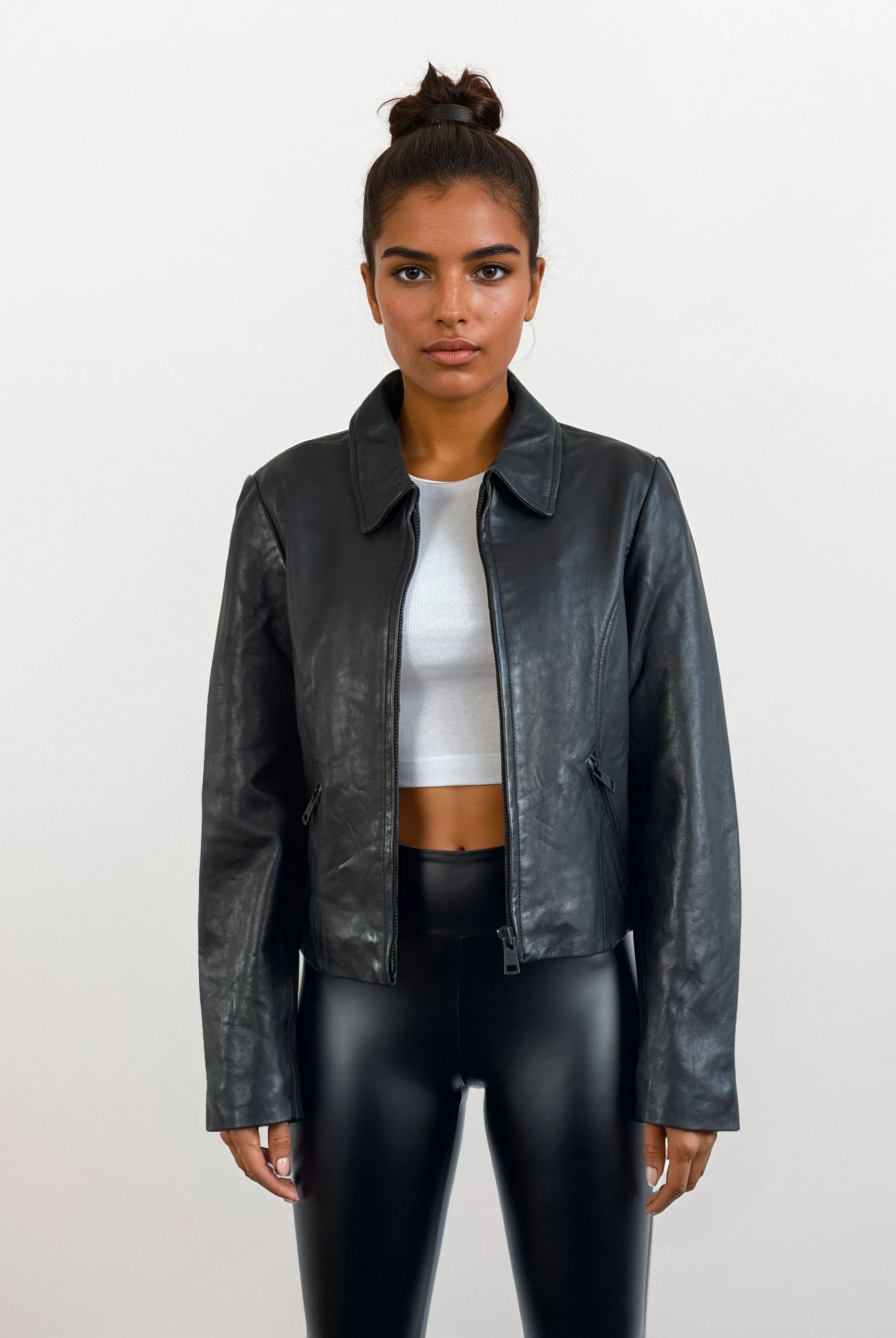 Eine Person trägt die trueprodigy Damen Lederjacke aus Schafsleder in Schwarz mit minimalistischem Design über einem weißen Crop Top und glänzenden schwarzen Leggings vor einem schlichten, hellen Hintergrund.