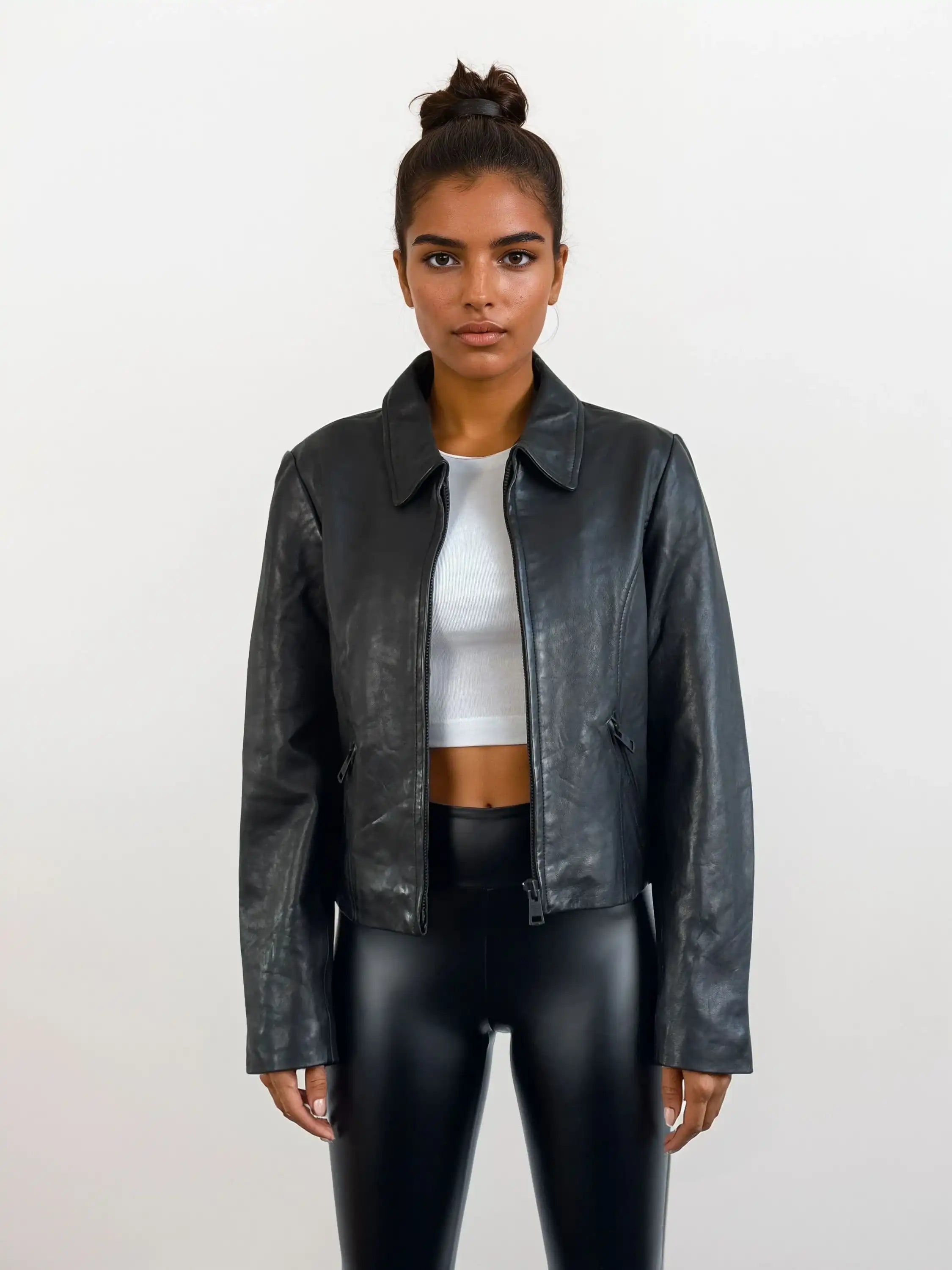 Eine Person trägt die trueprodigy Damen Lederjacke aus Schafsleder in Schwarz mit minimalistischem Design über einem weißen Crop Top und glänzenden schwarzen Leggings vor einem schlichten, hellen Hintergrund.