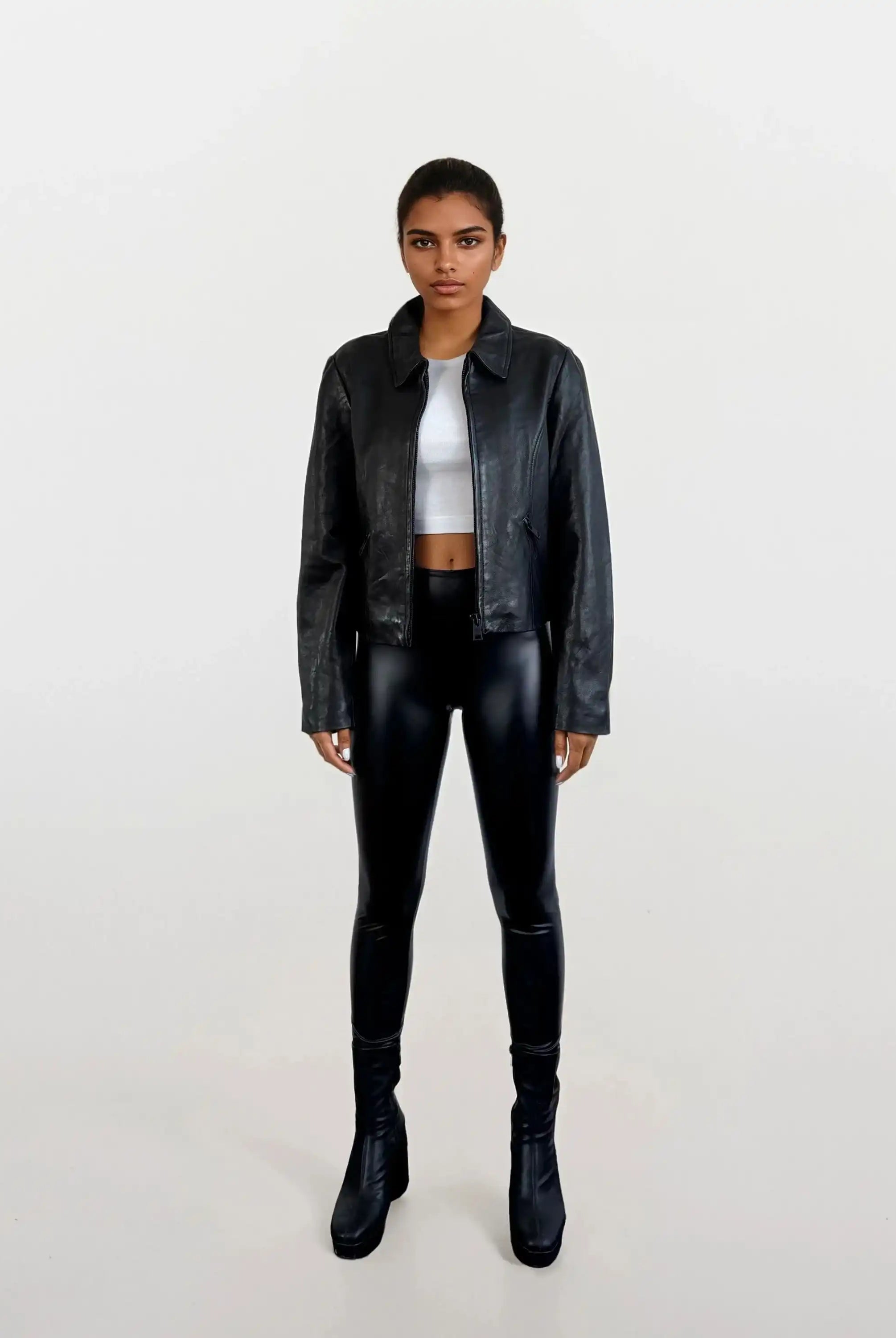 Eine Person steht mit dem Gesicht nach vorne vor einem schlichten weißen Hintergrund und trägt die schwarze Damen Lederjacke aus Schafsleder mit minimalistischem Design von trueprodigy, schwarze glänzende Leggings, ein weißes Crop-Top und schwarze hochhackige Stiefel.