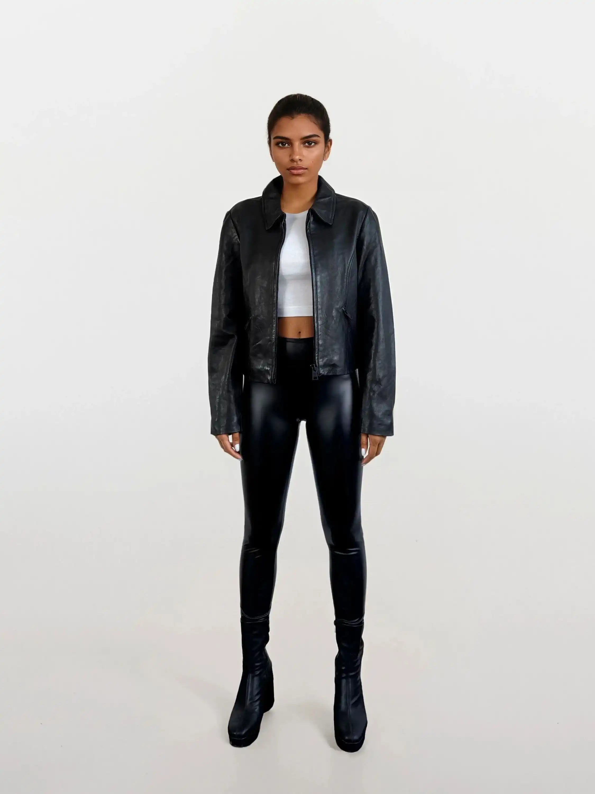 Eine Person steht mit dem Gesicht nach vorne vor einem schlichten weißen Hintergrund und trägt die schwarze Damen Lederjacke aus Schafsleder mit minimalistischem Design von trueprodigy, schwarze glänzende Leggings, ein weißes Crop-Top und schwarze hochhackige Stiefel.