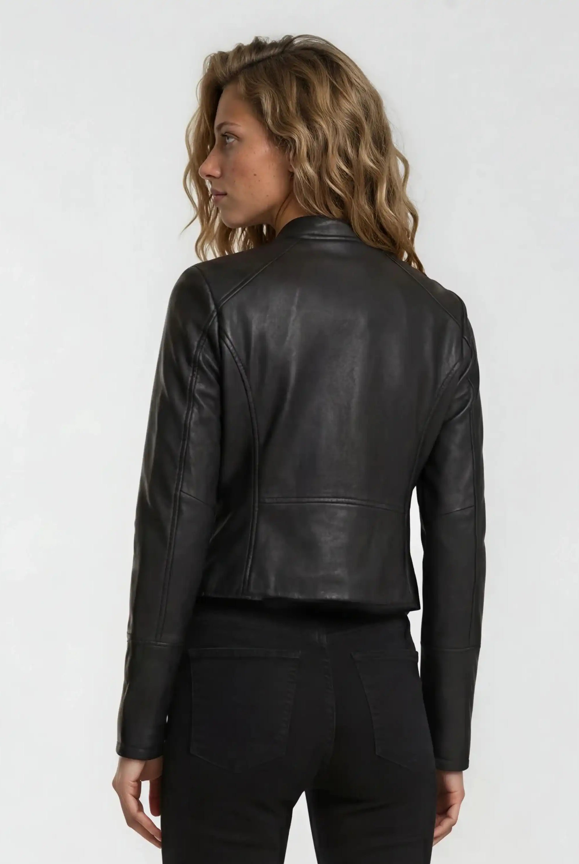 Eine Person mit gewelltem, hellbraunem Haar trägt die trueprodigy Damen Lederjacke aus Schafsleder in Schwarz im sportlichen Racing-Stil und eine schwarze Hose und blickt von der Kamera weg auf einen schlichten, hellen Hintergrund.