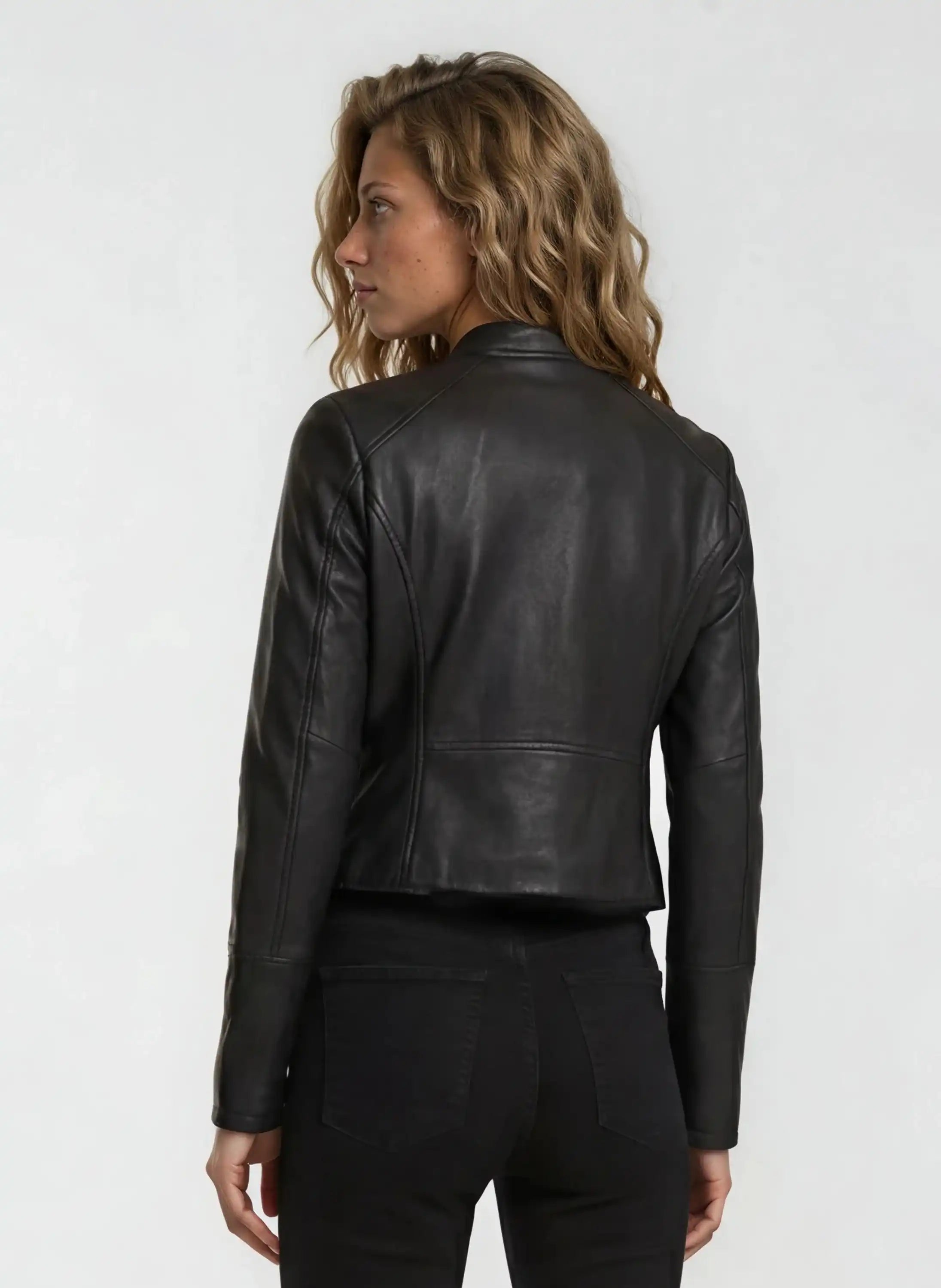 Eine Person mit gewelltem, hellbraunem Haar trägt die trueprodigy Damen Lederjacke aus Schafsleder in Schwarz im sportlichen Racing-Stil und eine schwarze Hose und blickt von der Kamera weg auf einen schlichten, hellen Hintergrund.