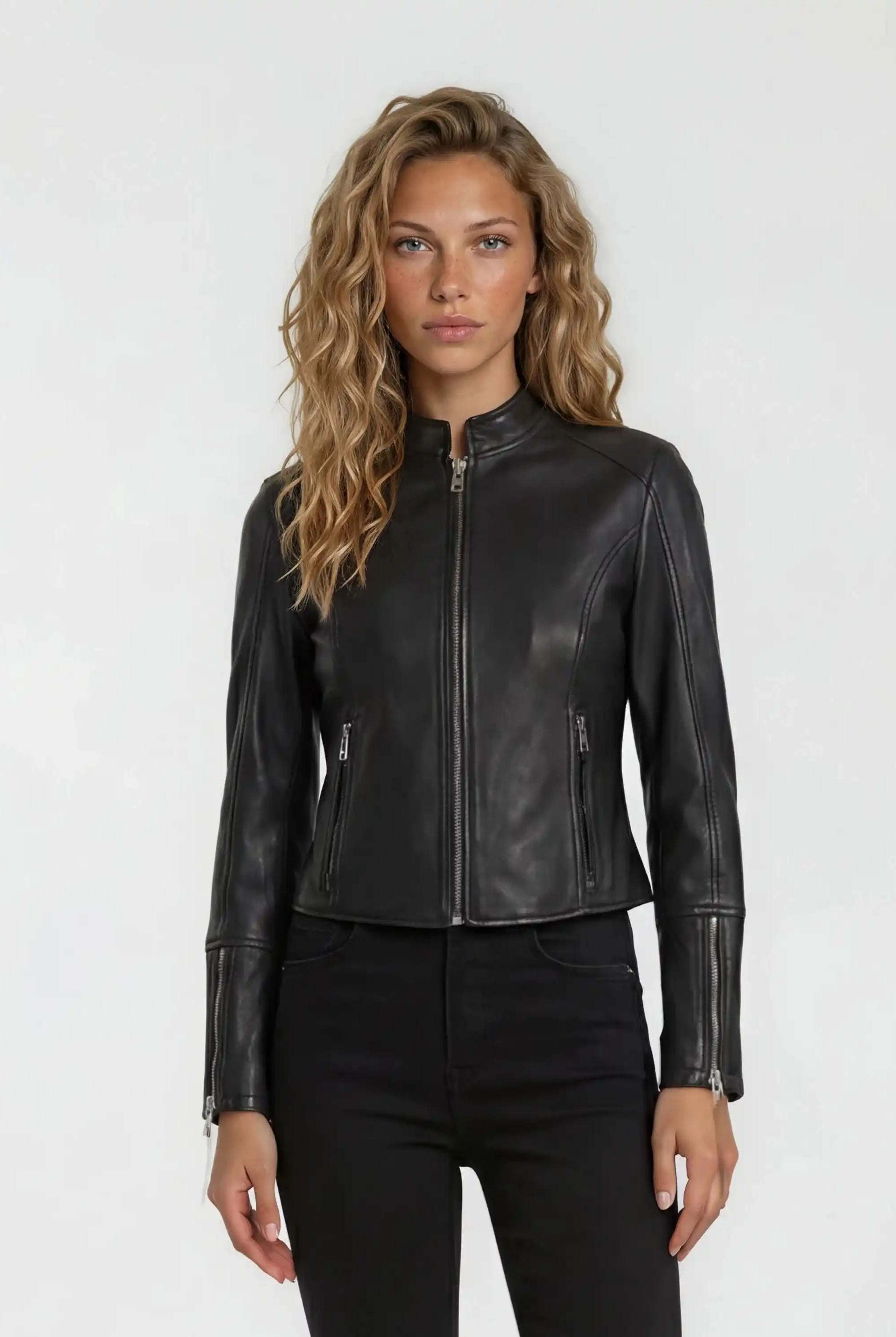 Eine Person mit gewelltem, hellbraunem Haar steht vor einem schlichten Hintergrund und trägt die trueprodigy Damen Lederjacke aus Schafsleder in Schwarz im sportlichen Racing-Stil, gepaart mit einer schwarzen Hose.