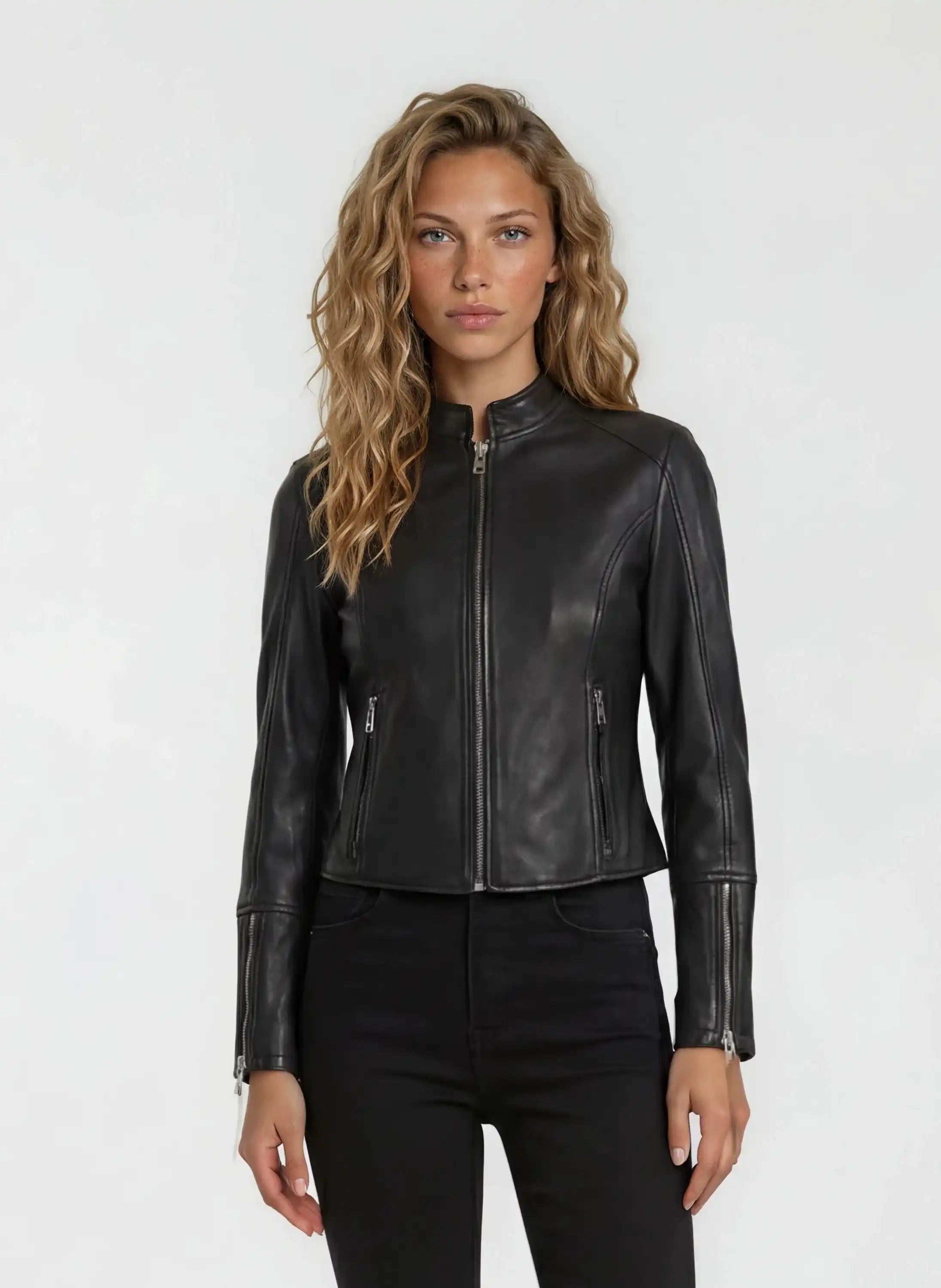 Eine Person mit gewelltem, hellbraunem Haar steht vor einem schlichten Hintergrund und trägt die trueprodigy Damen Lederjacke aus Schafsleder in Schwarz im sportlichen Racing-Stil, gepaart mit einer schwarzen Hose.