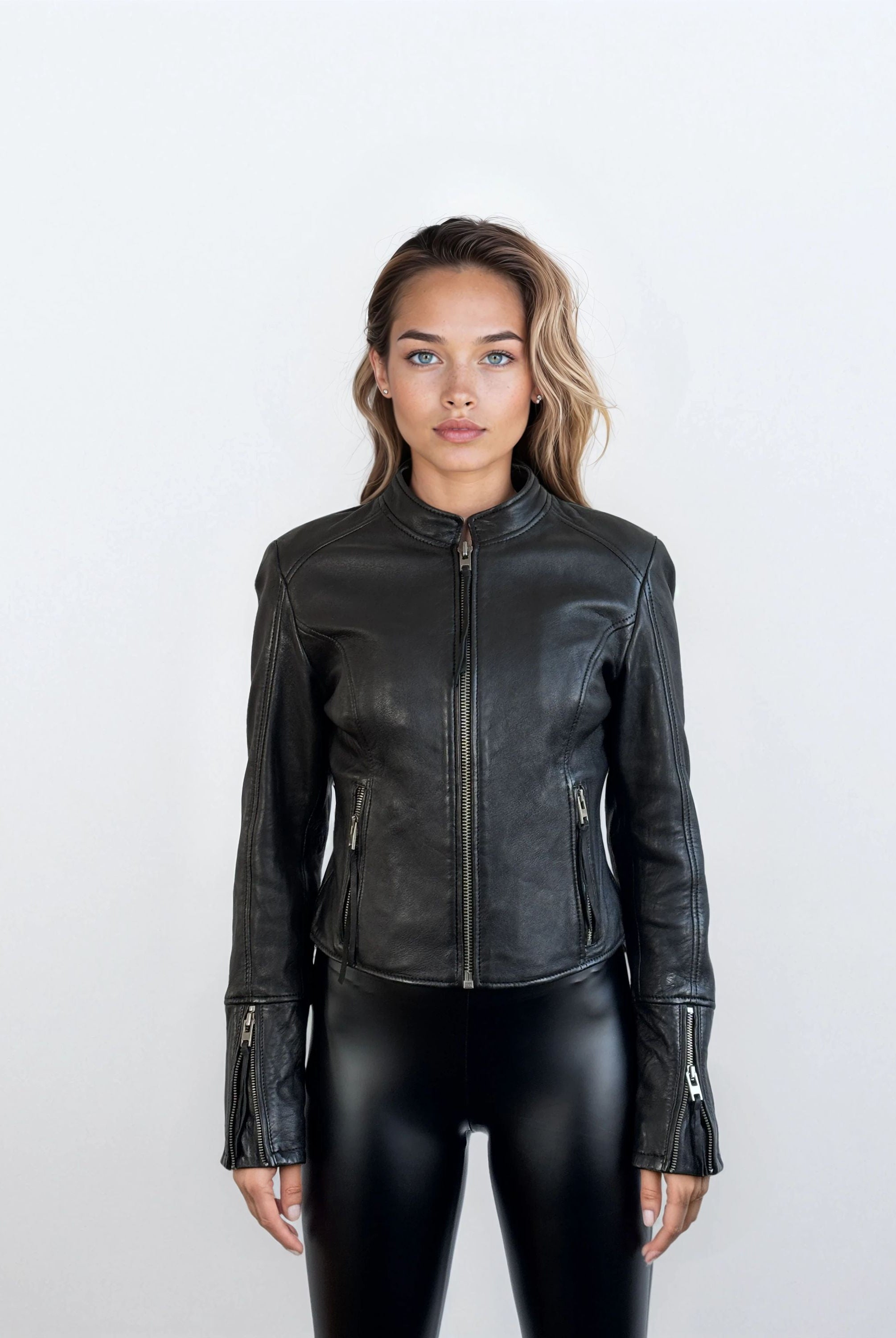 Eine Frau mit offenem Haar und neutralem Gesichtsausdruck steht vor einem schlichten hellen Hintergrund vor der Kamera. Sie trägt die trueprodigy Damen Lederjacke aus Schafsleder in Schwarz im sportlichen Racing-Stil und eine glänzende schwarze Hose.