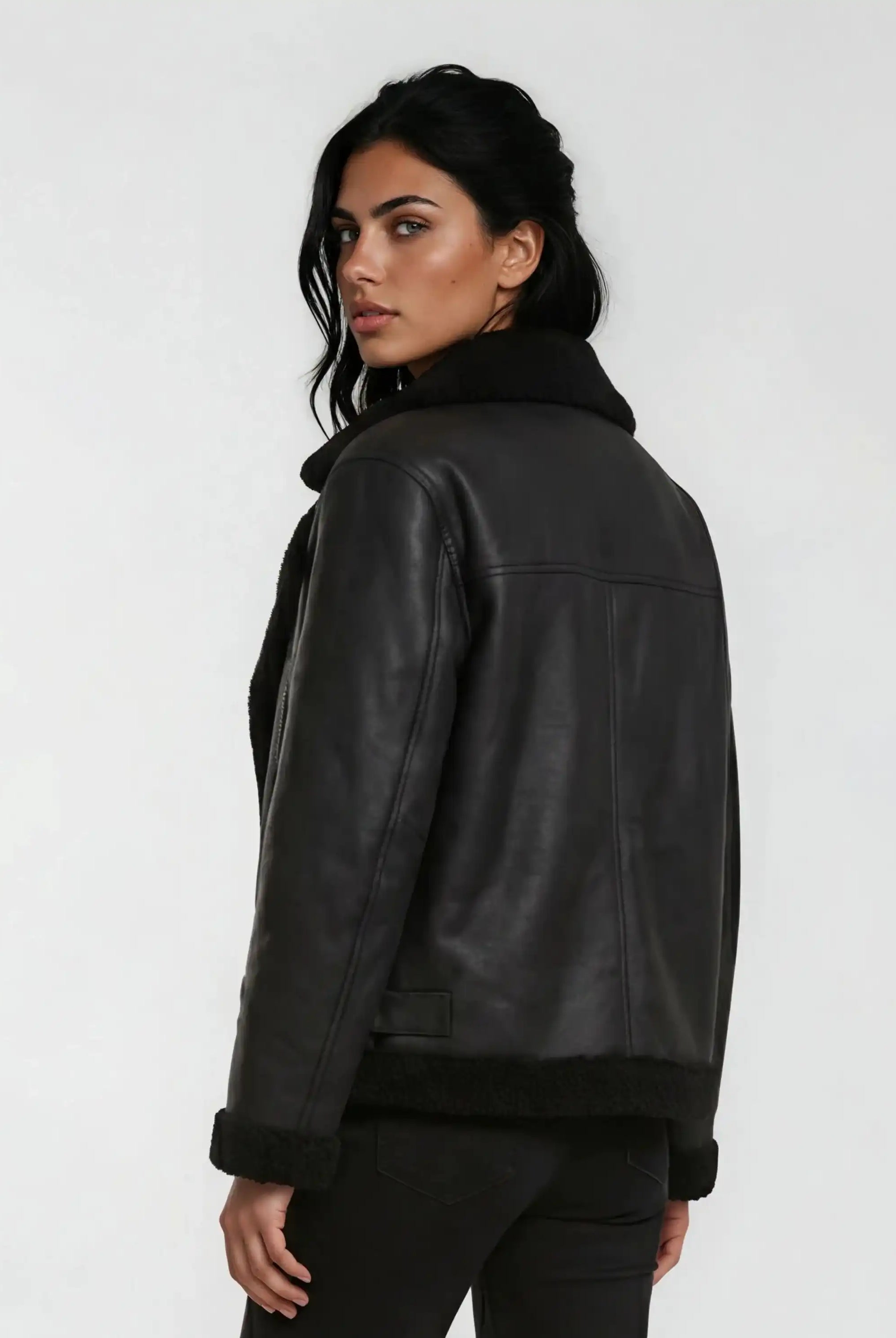 Eine Person mit langen dunklen Haaren in lockeren Wellen trägt die trueprodigy Damen Lederjacke aus Schafsleder in Schwarz mit Teddy-Besatz, steht vor einem schlichten hellgrauen Hintergrund und schaut über ihre Schulter.