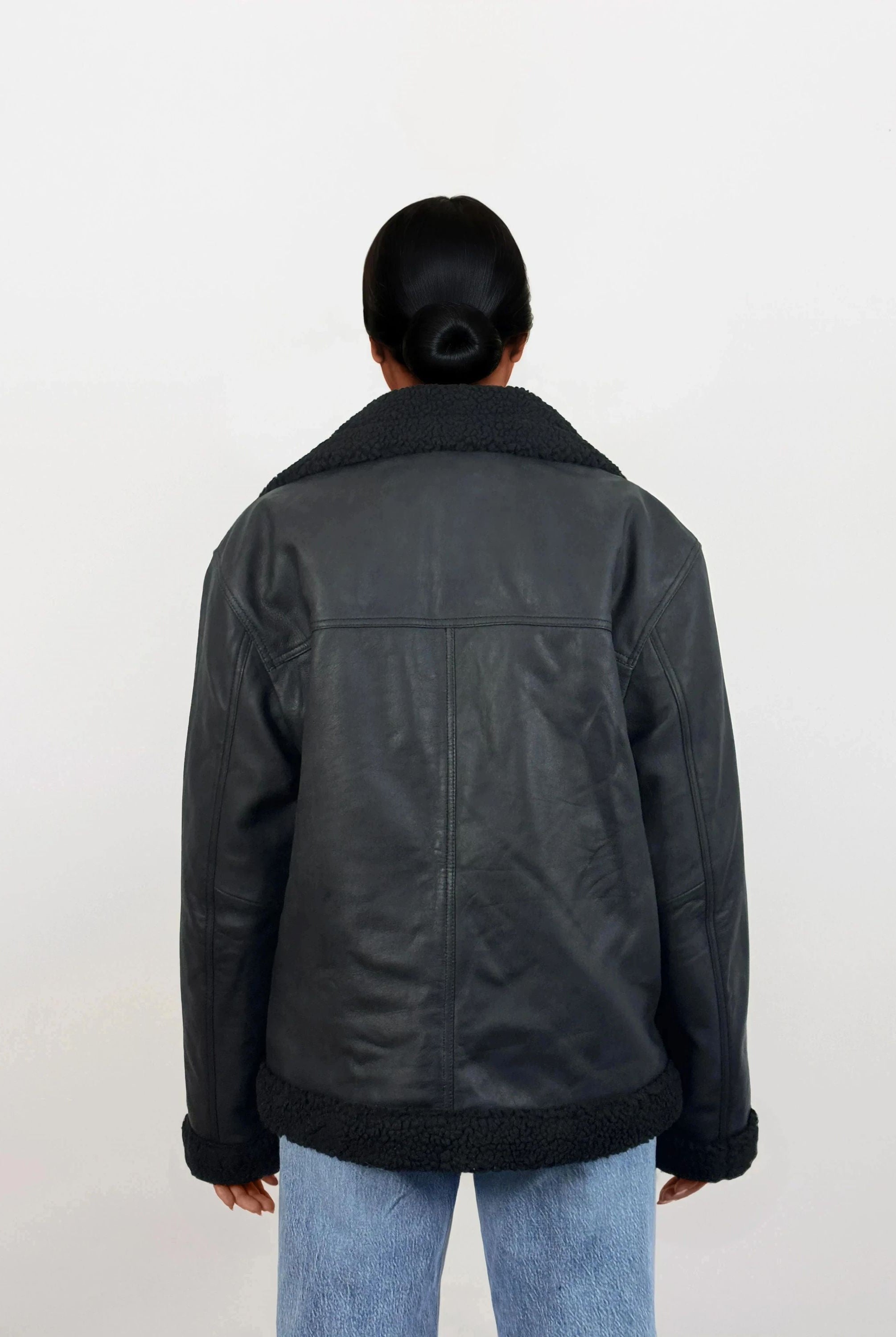 Eine Person mit dunklen Haaren in einem tiefen Dutt wird von hinten gezeigt. Sie trägt eine trueprodigy Damen Lederjacke aus Schafsleder in Schwarz mit Teddy-Besatz und hellblaue Jeans vor einem schlichten weißen Hintergrund.