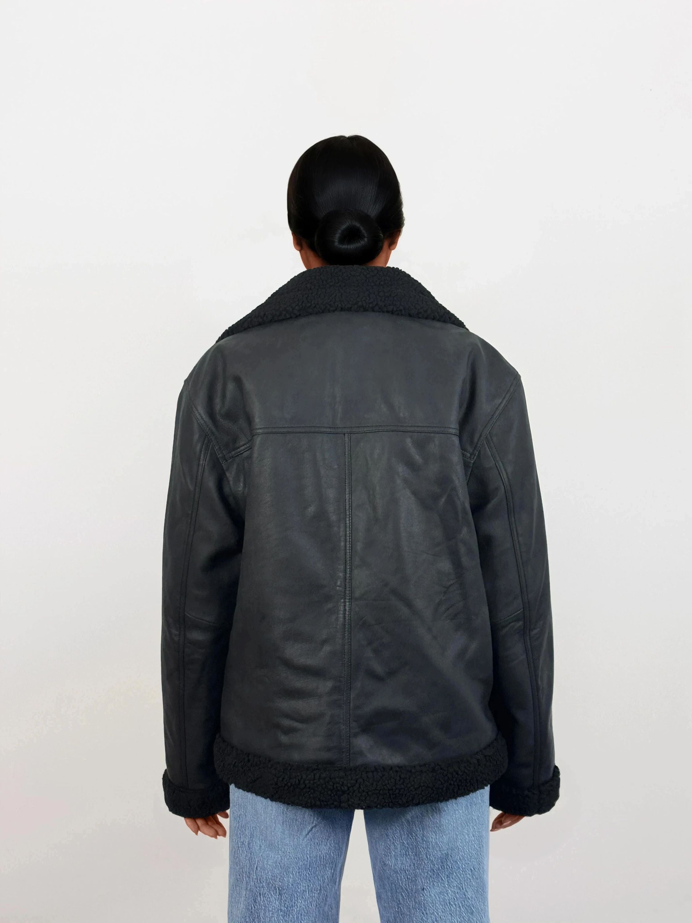 Eine Person mit dunklen Haaren in einem tiefen Dutt wird von hinten gezeigt. Sie trägt eine trueprodigy Damen Lederjacke aus Schafsleder in Schwarz mit Teddy-Besatz und hellblaue Jeans vor einem schlichten weißen Hintergrund.