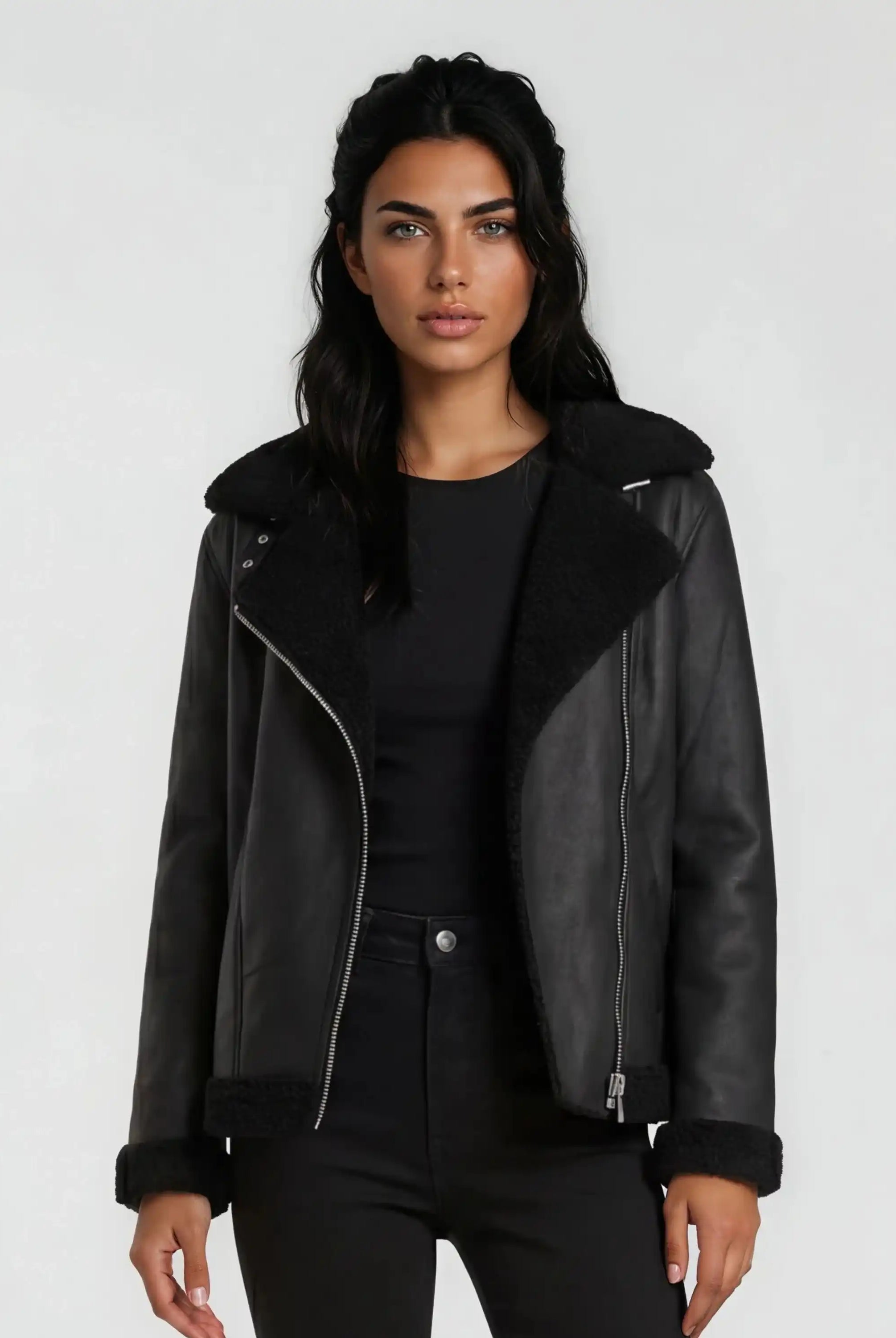 Eine Frau mit langen dunklen Haaren modelliert die trueprodigy Damen Lederjacke aus Schafsleder in Schwarz mit Teddy-Besatz über einem schwarzen Oberteil und einer Hose vor einem schlichten hellgrauen Hintergrund.