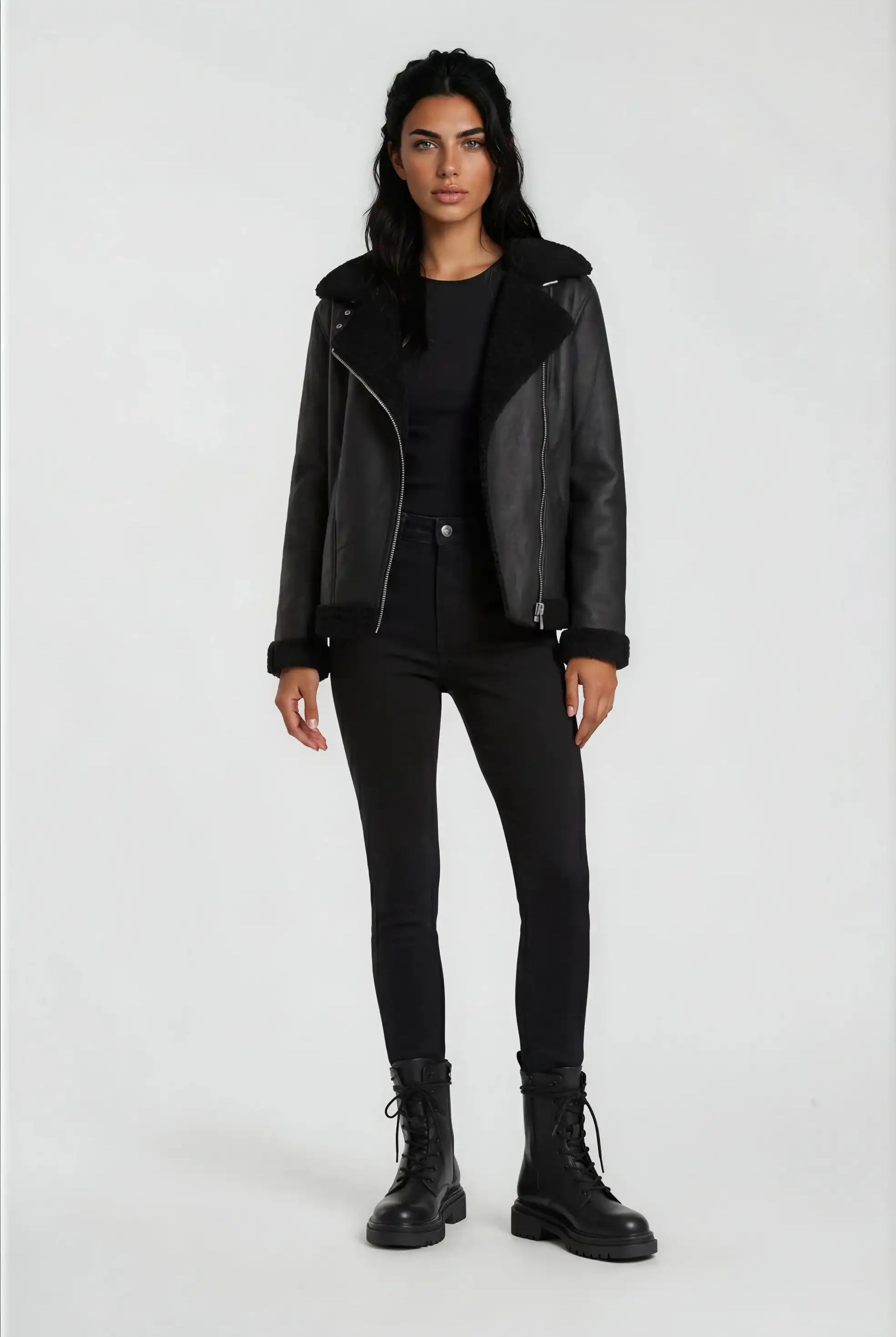 Eine Frau steht mit dem Gesicht nach vorne vor einem hellen Hintergrund und trägt die trueprodigy Damen Lederjacke aus Schafsleder in Schwarz mit Teddy-Besatz, gepaart mit einem schwarzen Top, Skinny Jeans und Schnürstiefeln. Ihre Arme ruhen an den Seiten.