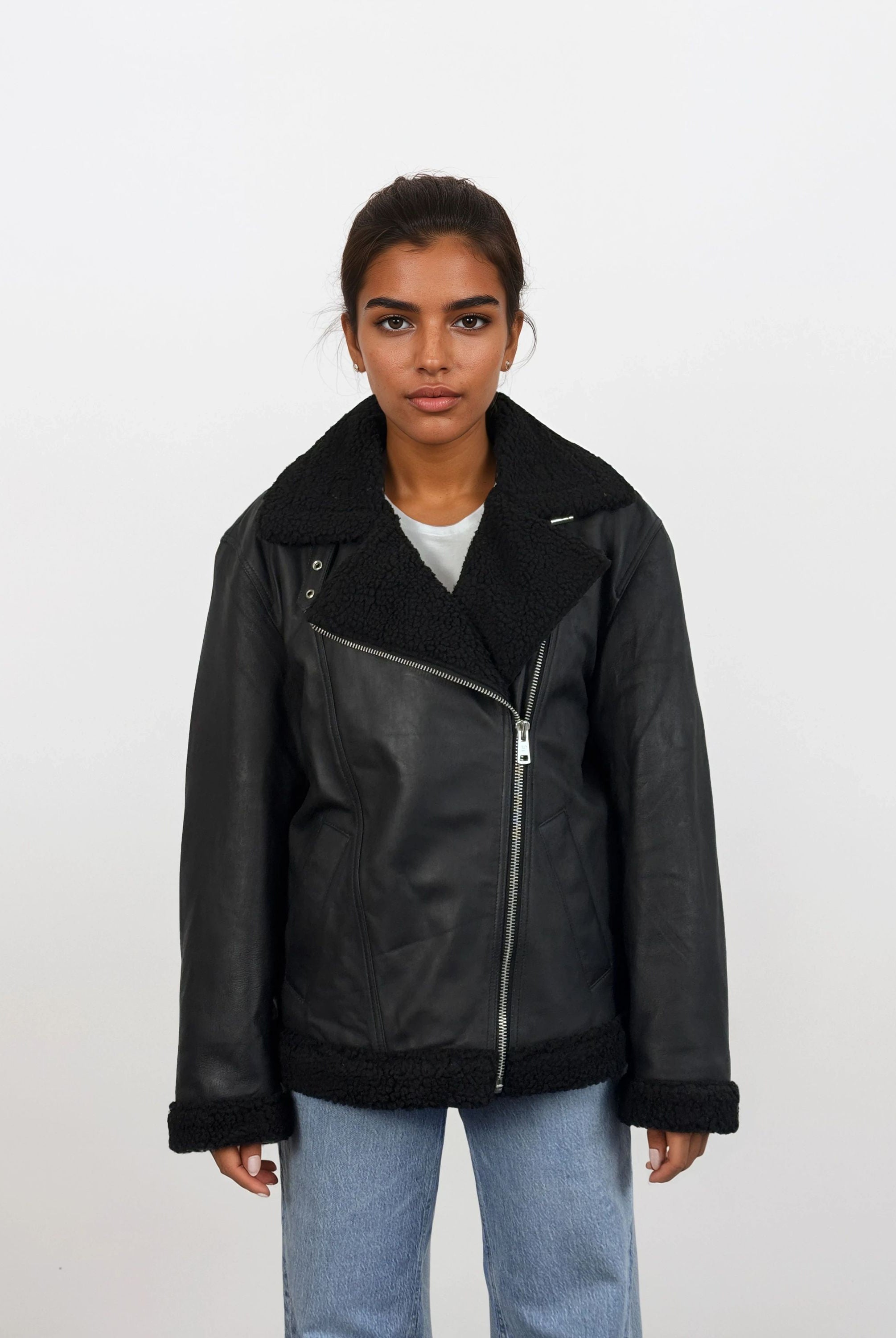 Eine Frau mit dunklem, nach hinten gebundenem Haar trägt die trueprodigy Damen Lederjacke aus Schafsleder in Schwarz mit Teddy-Besatz über einem weißen Hemd und hellblauen Jeans, vor einem schlichten weißen Hintergrund.