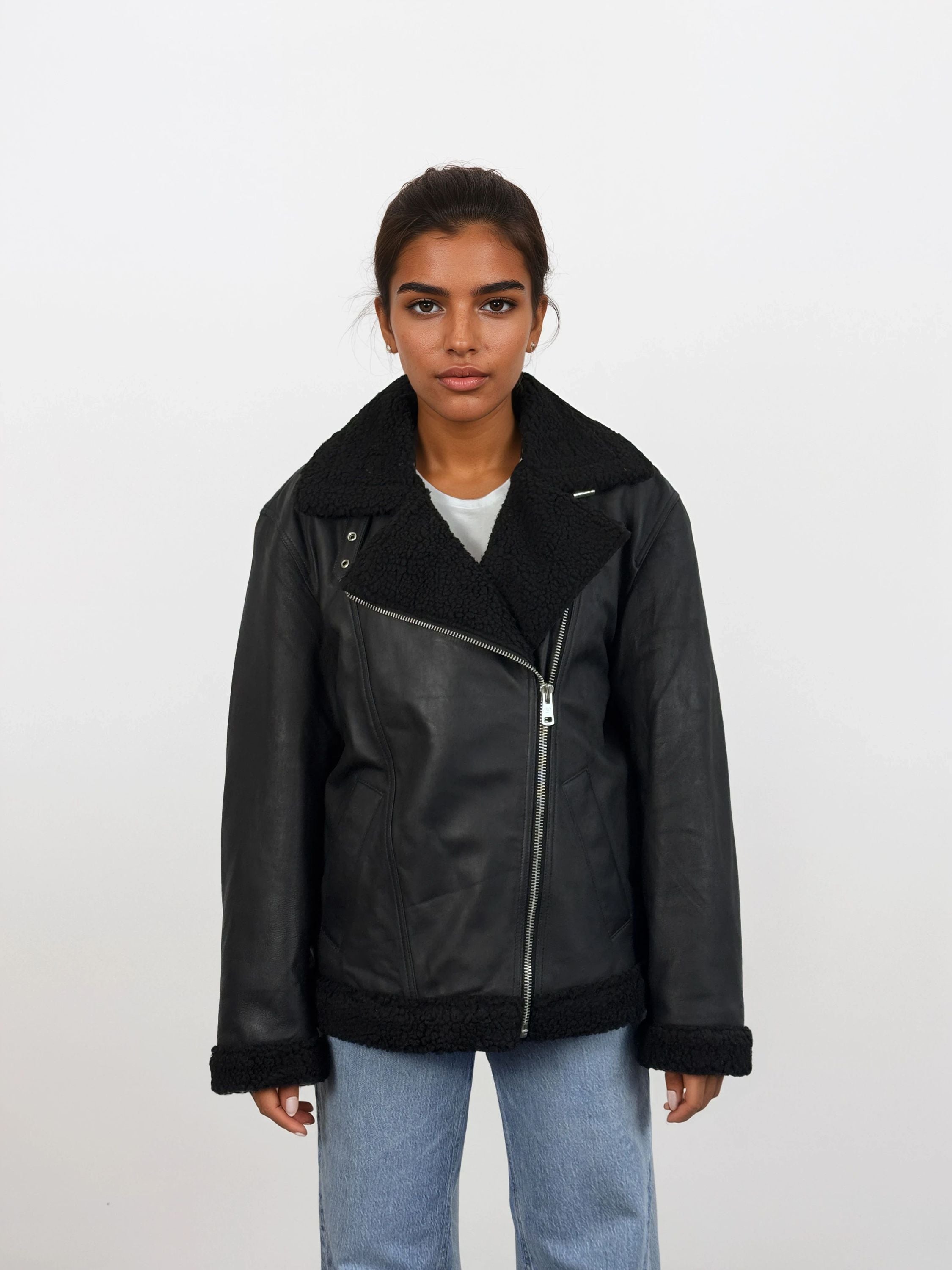 Eine Frau mit dunklem, nach hinten gebundenem Haar trägt die trueprodigy Damen Lederjacke aus Schafsleder in Schwarz mit Teddy-Besatz über einem weißen Hemd und hellblauen Jeans, vor einem schlichten weißen Hintergrund.
